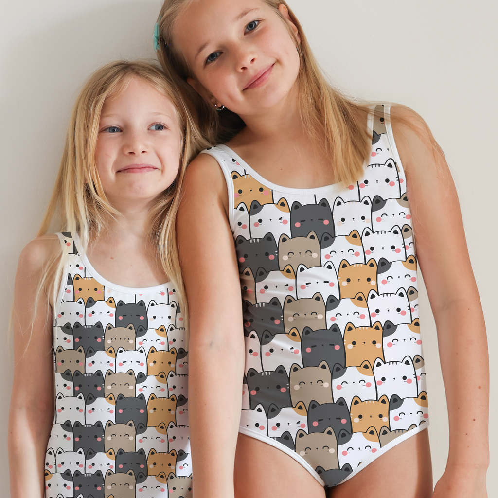 Two young girls wearing matching white one-piece swimsuits with a playful cartoon bear pattern, holding hands against a plain light gray background.