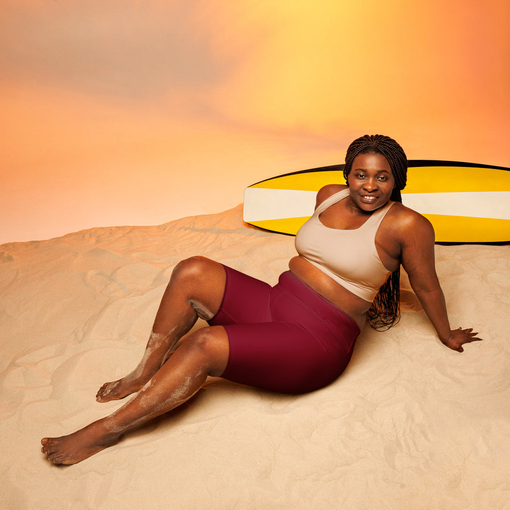 A woman lounges on sand wearing women’s burgundy high waisted moisture wicking biker shorts next to a yellow surfboard