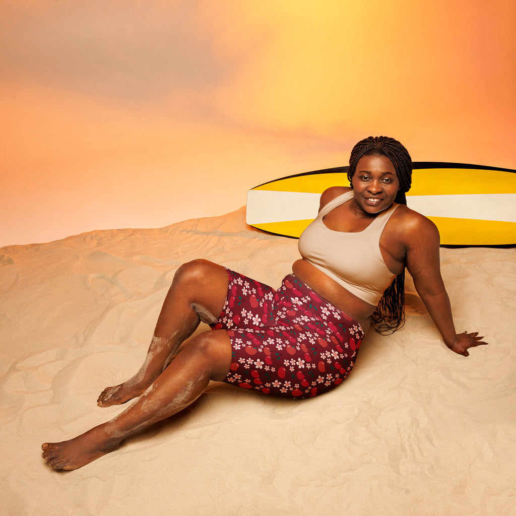 A woman wearing women’s burgundy cherry daisy high waisted moisture wicking biker shorts sitting on sand next to a surfboard