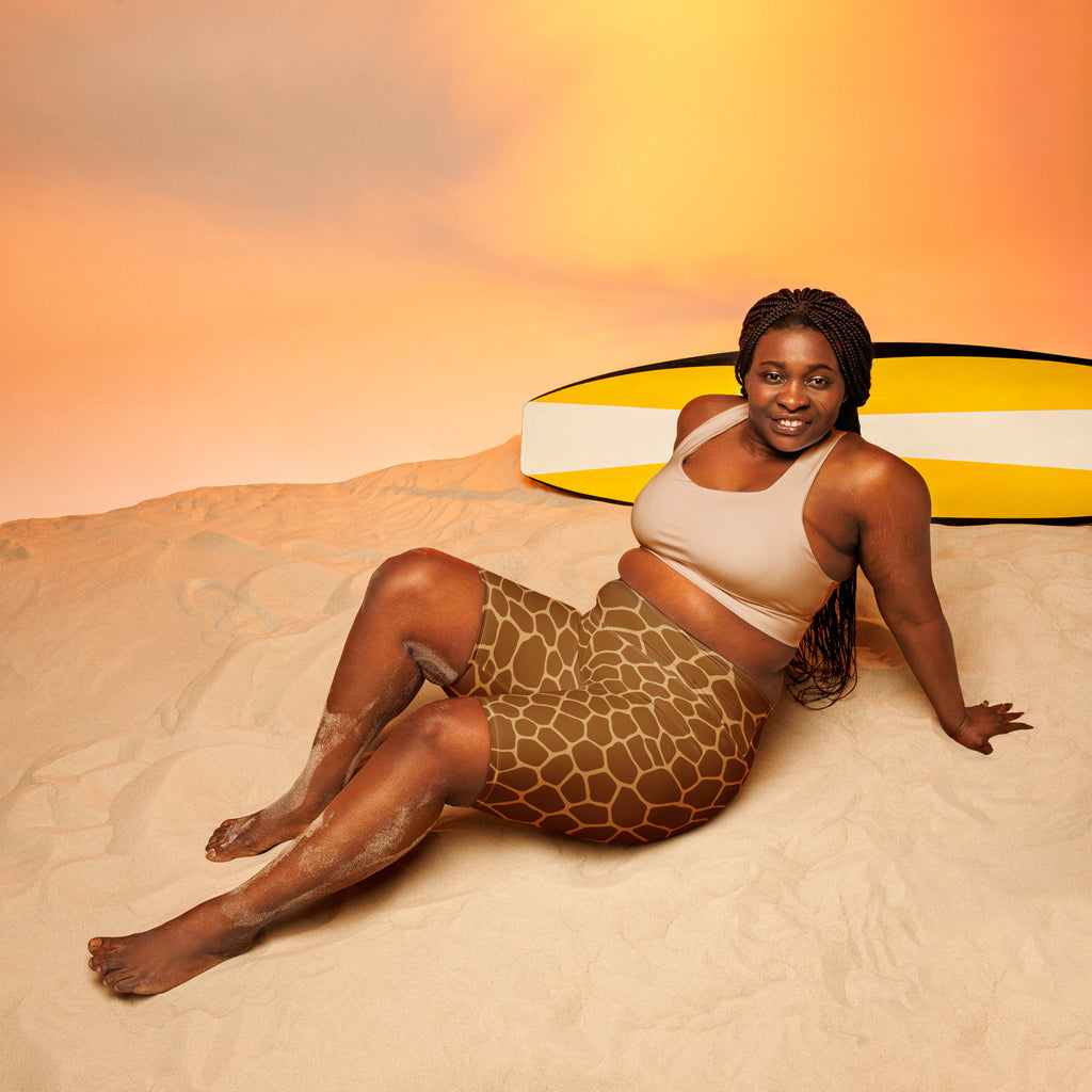 A woman wearing beige tank top and brown giraffe print moisture wicking high waisted biker shorts reclining on sand