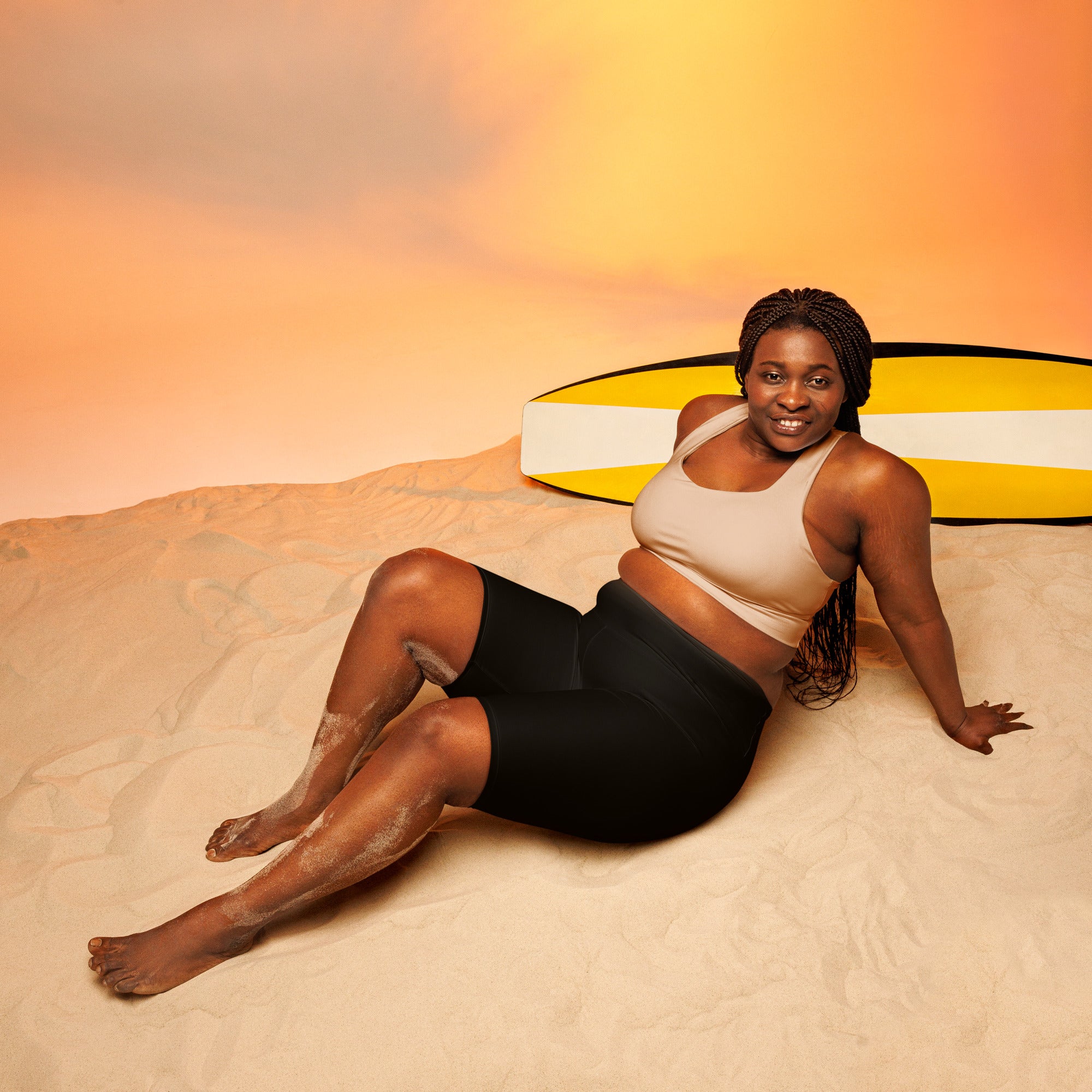 A woman lounges on sand wearing black high waisted moisture wicking biker shorts with a yellow surfboard nearby