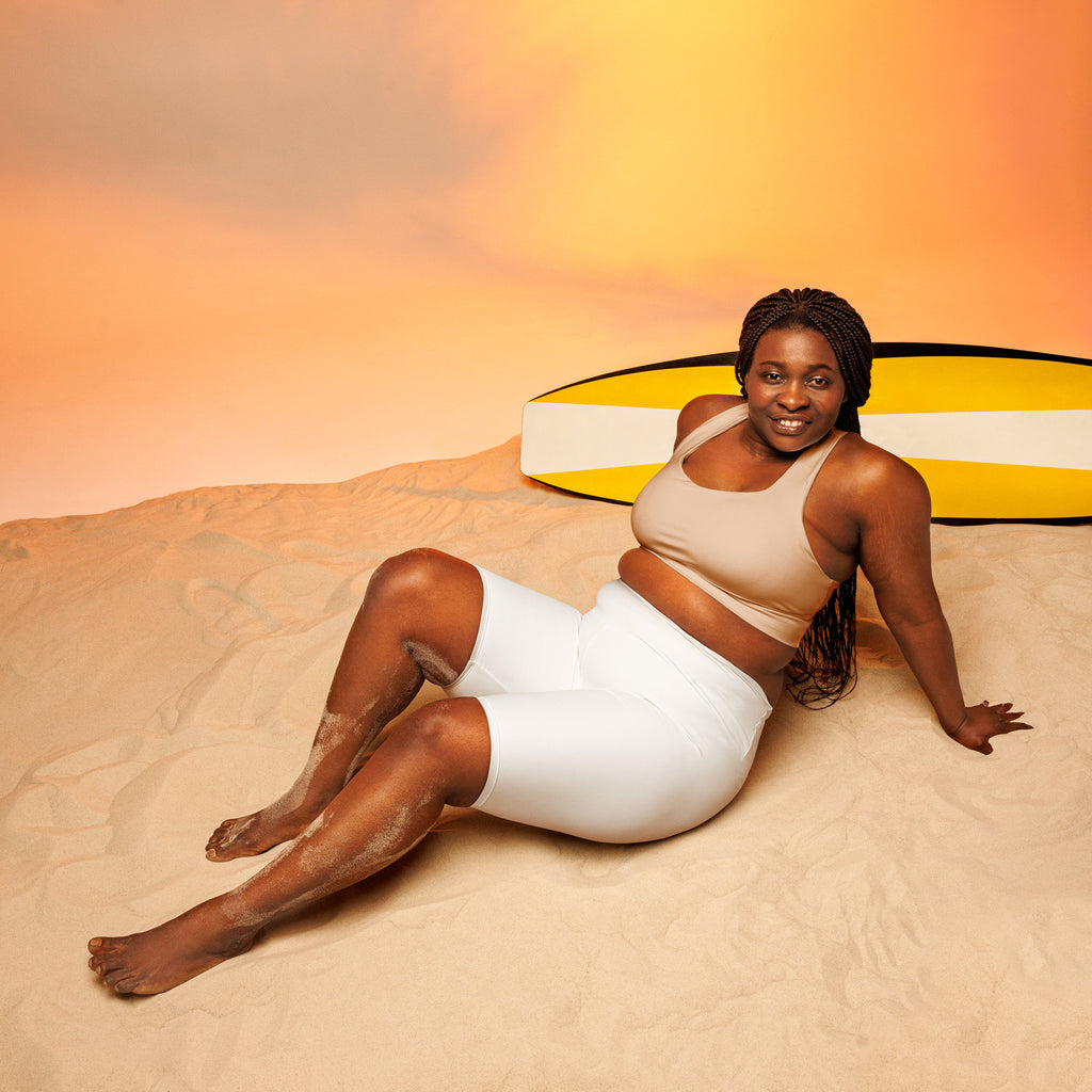 A woman lounges on sand wearing women’s white high waisted moisture wicking biker shorts with a yellow surfboard
