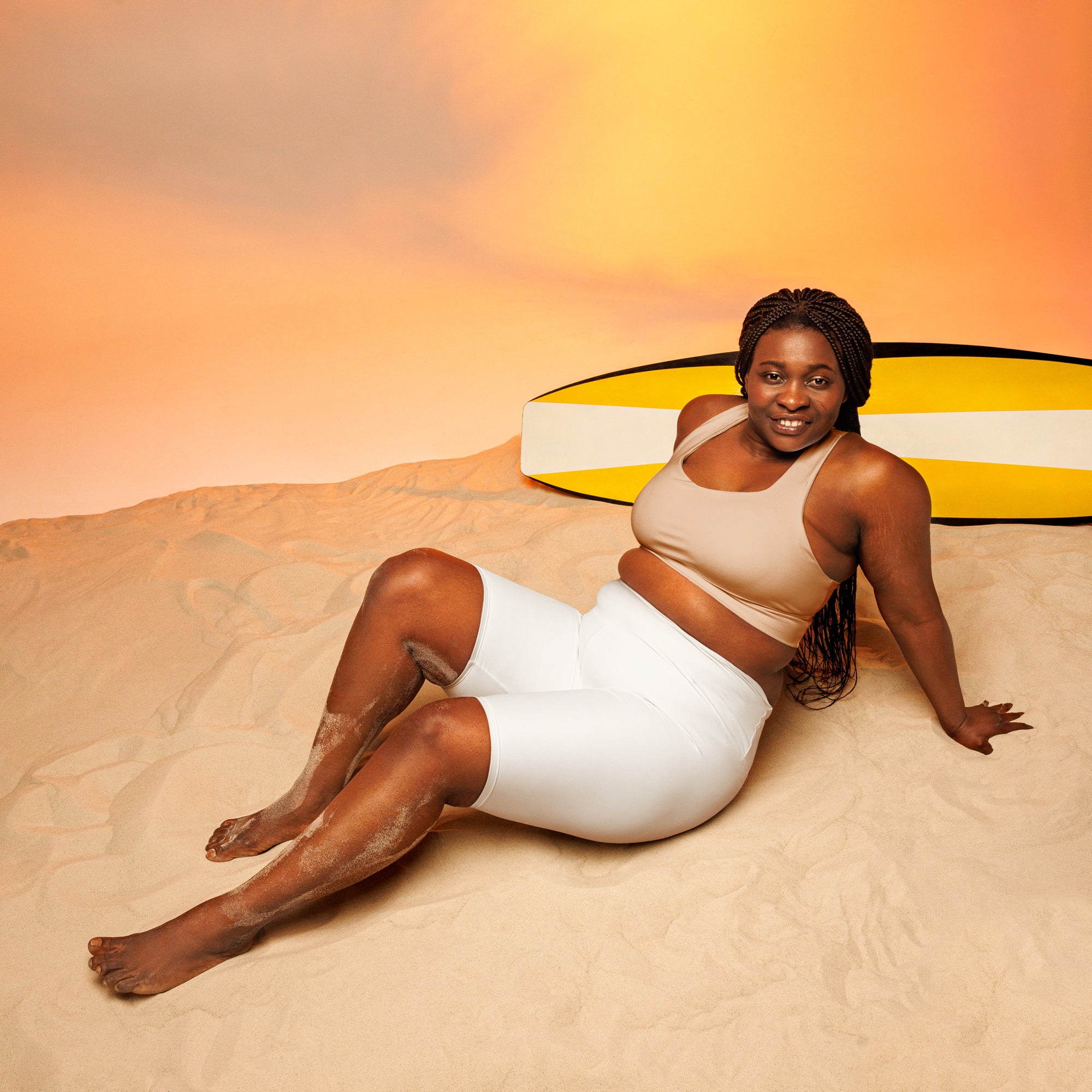 A woman lounges on sand wearing women’s white high waisted moisture wicking biker shorts with a yellow surfboard