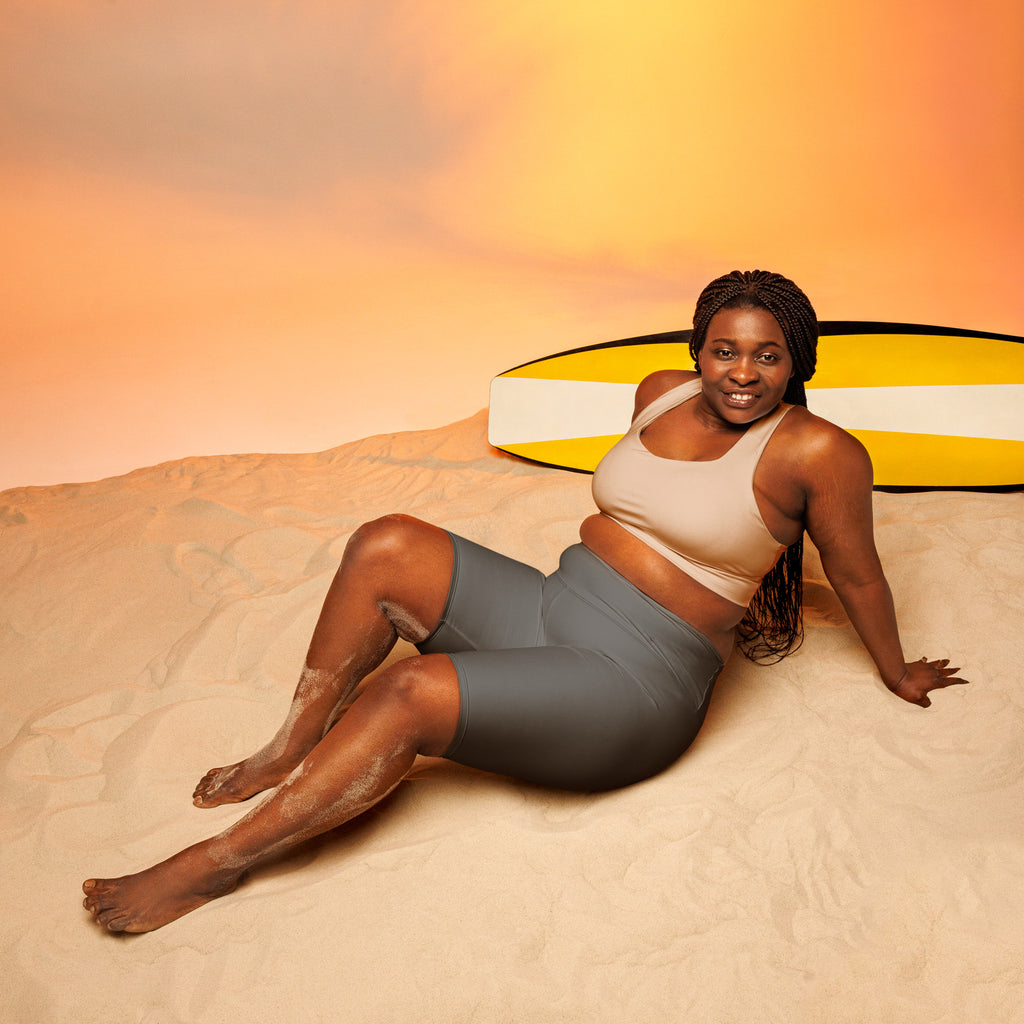 A woman lounges on sand wearing women’s gray high waisted moisture wicking biker shorts with a yellow surfboard behind her