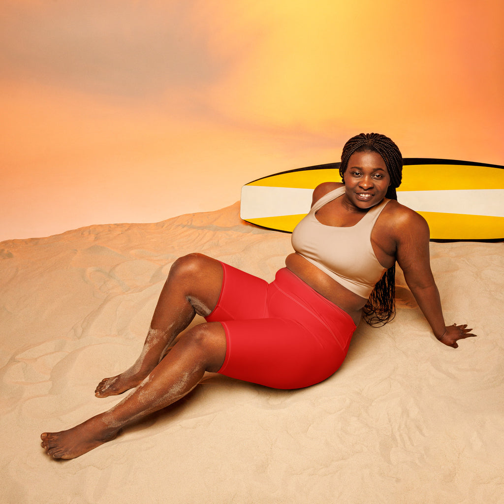 A woman wearing moisture-wicking women’s red high waisted biker shorts reclining on sand with a yellow surfboard behind her
