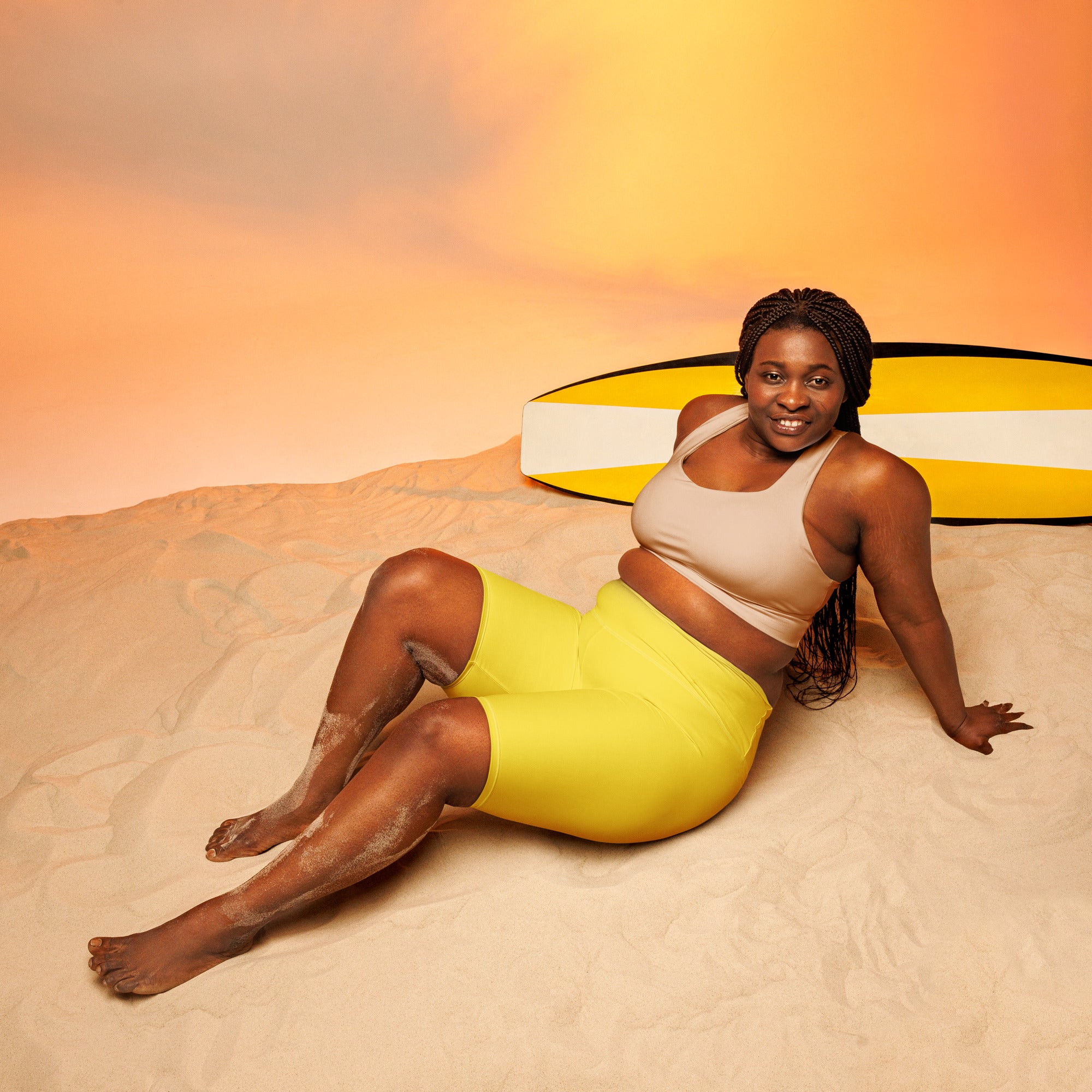 A woman lounges on sand wearing women’s yellow high waisted biker shorts moisture wicking active fit with a surfboard behind her