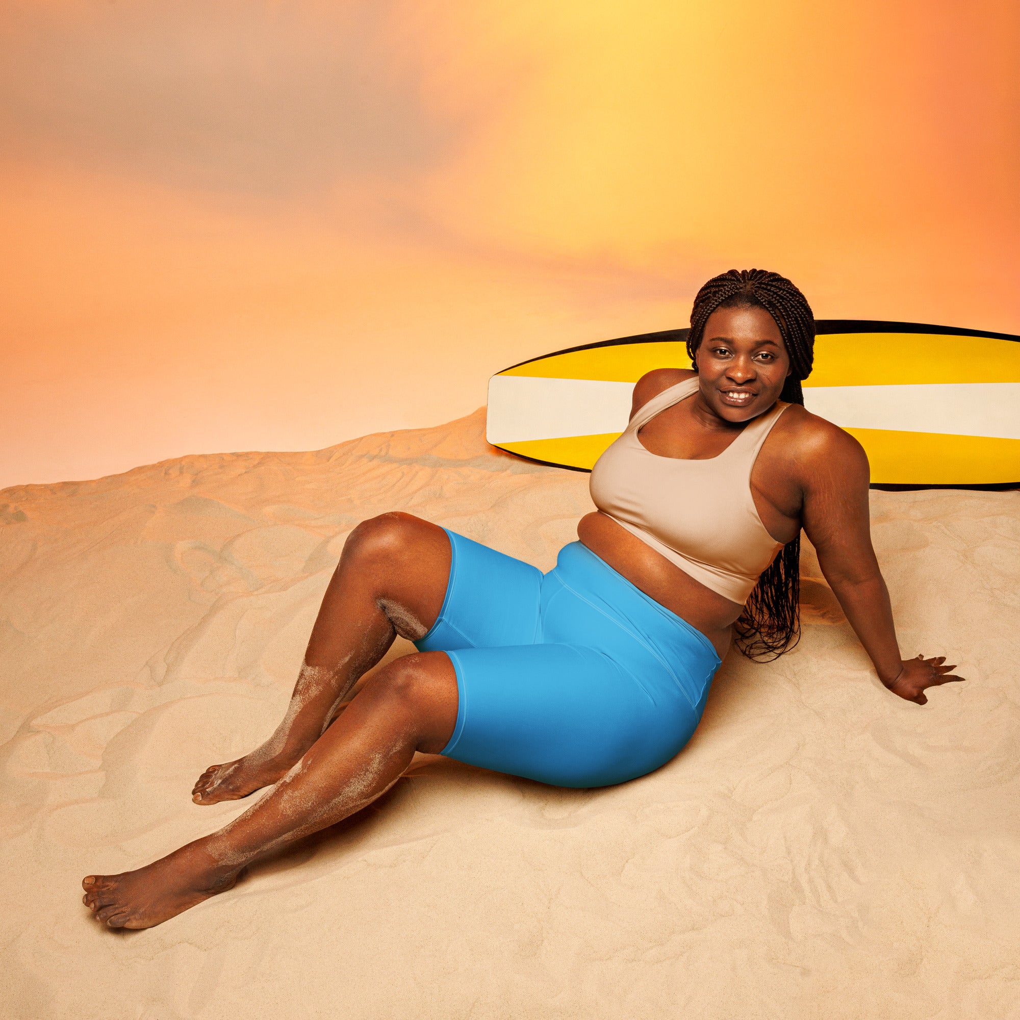 A woman lying on sand wearing sky blue high waisted moisture wicking biker shorts with a yellow surfboard behind her