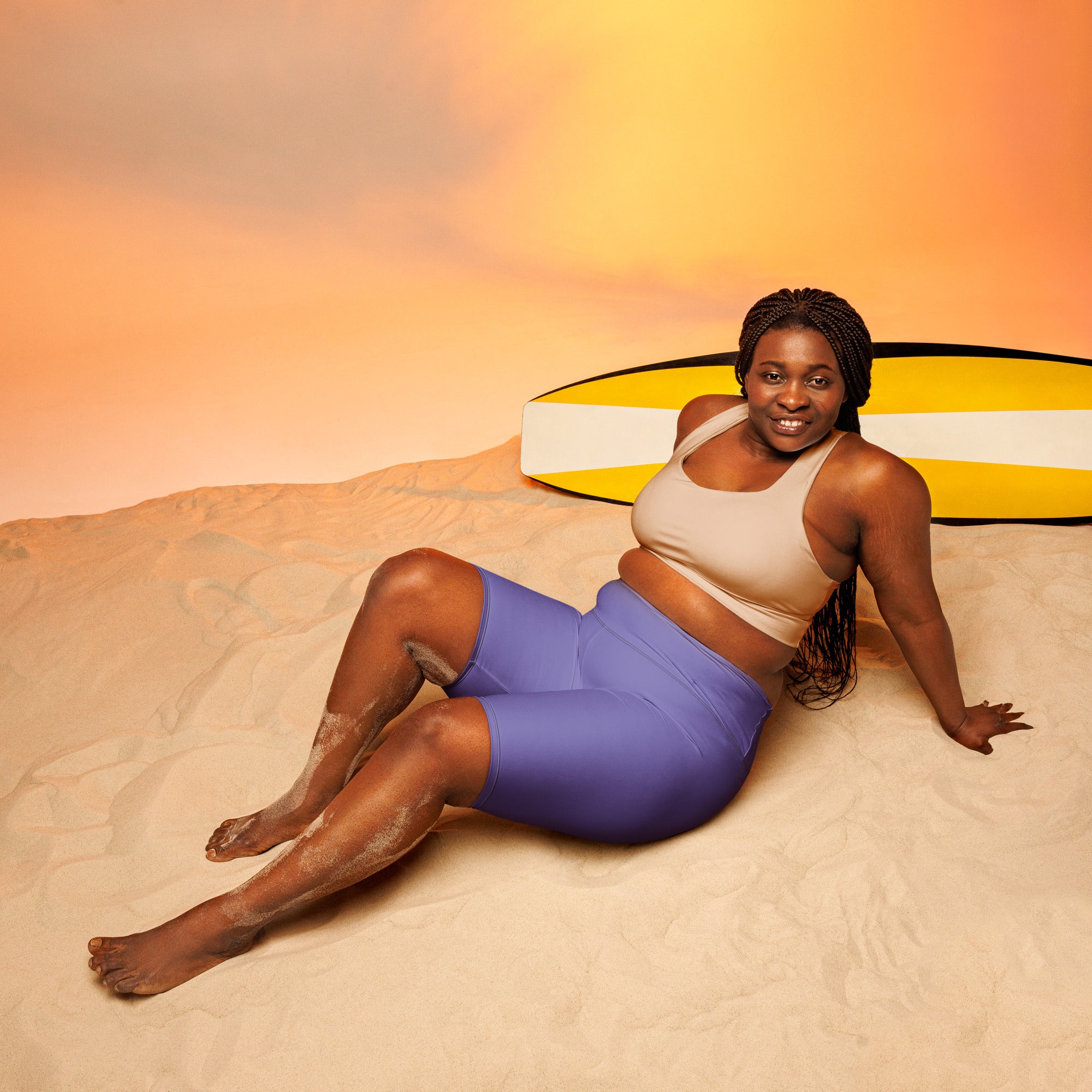 A woman lounges on sand wearing women’s purple high waisted moisture wicking biker shorts next to a surfboard