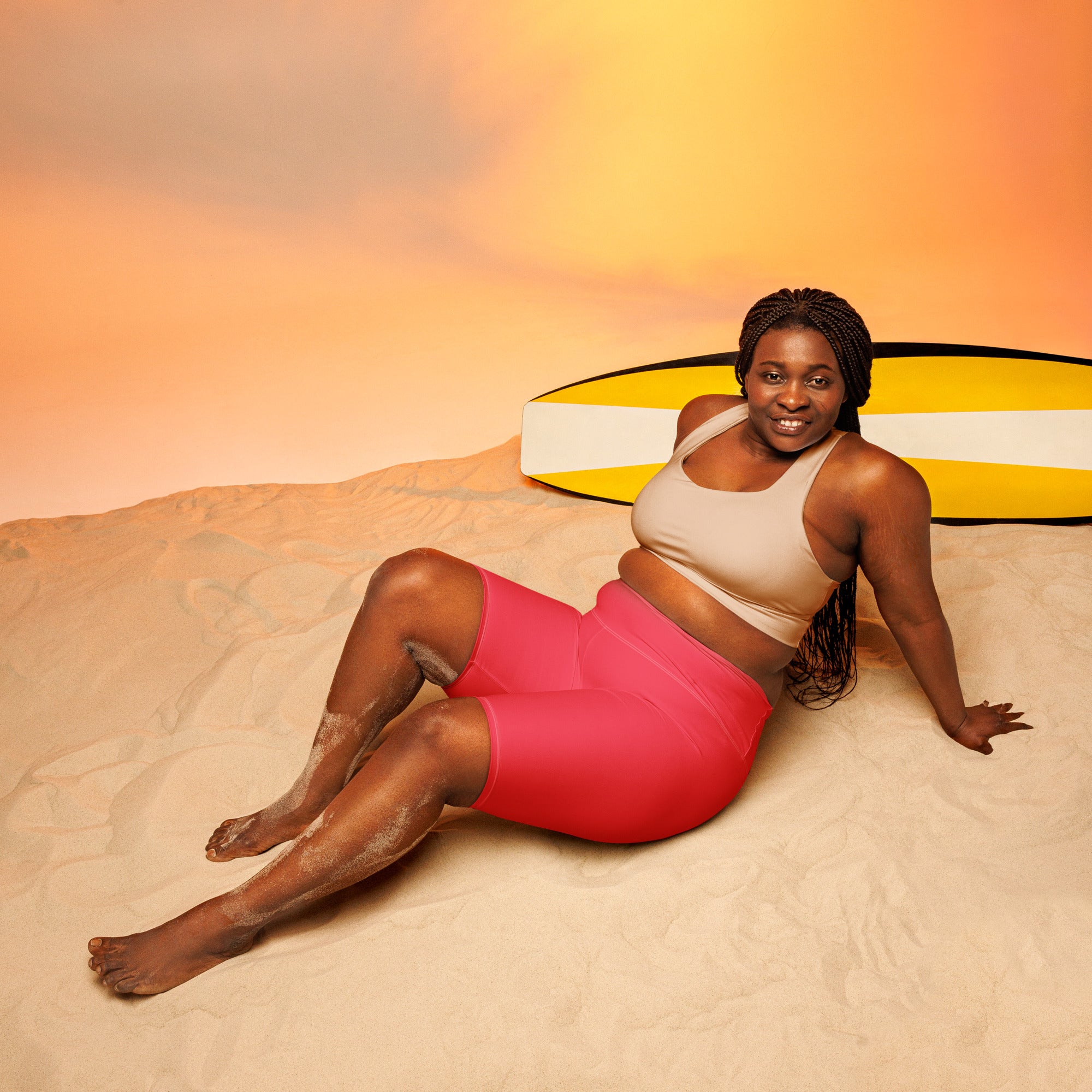 A woman lounges on sand wearing beige sports bra and bright pink high waisted moisture wicking biker shorts with surfboard