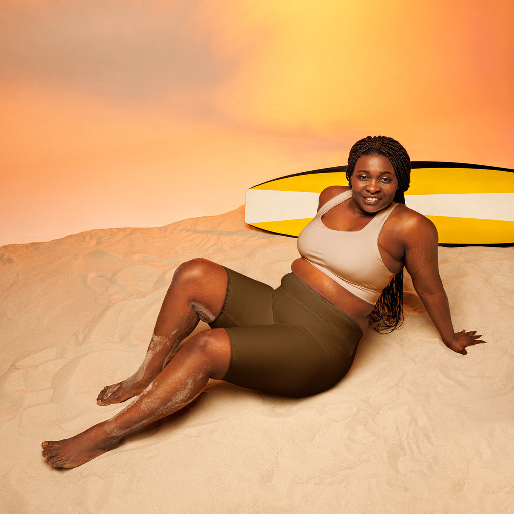 A woman lounges on sand wearing olive green moisture wicking high waisted biker shorts with a yellow surfboard behind her