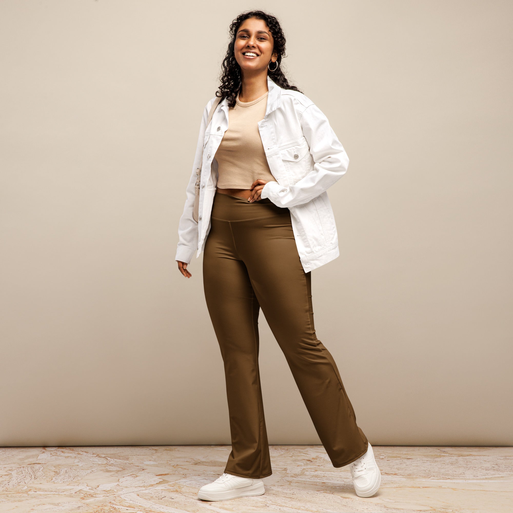 A woman wears brown high waist flare legging pants made of satin styled with a white denim jacket and white sneakers