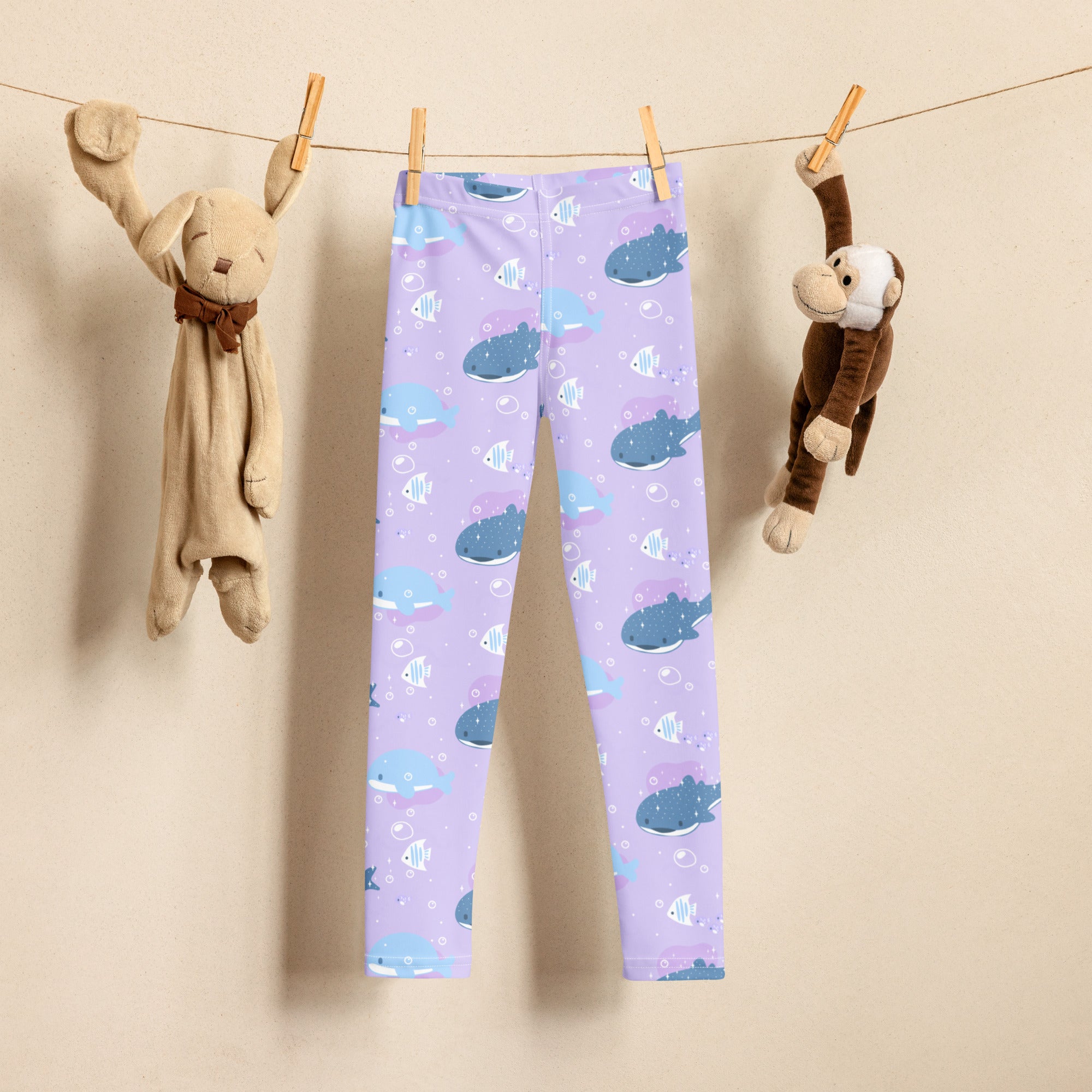 Girls purple buttery soft leggings with playful blue whale and small fish pattern hanging on clothesline between stuffed animals