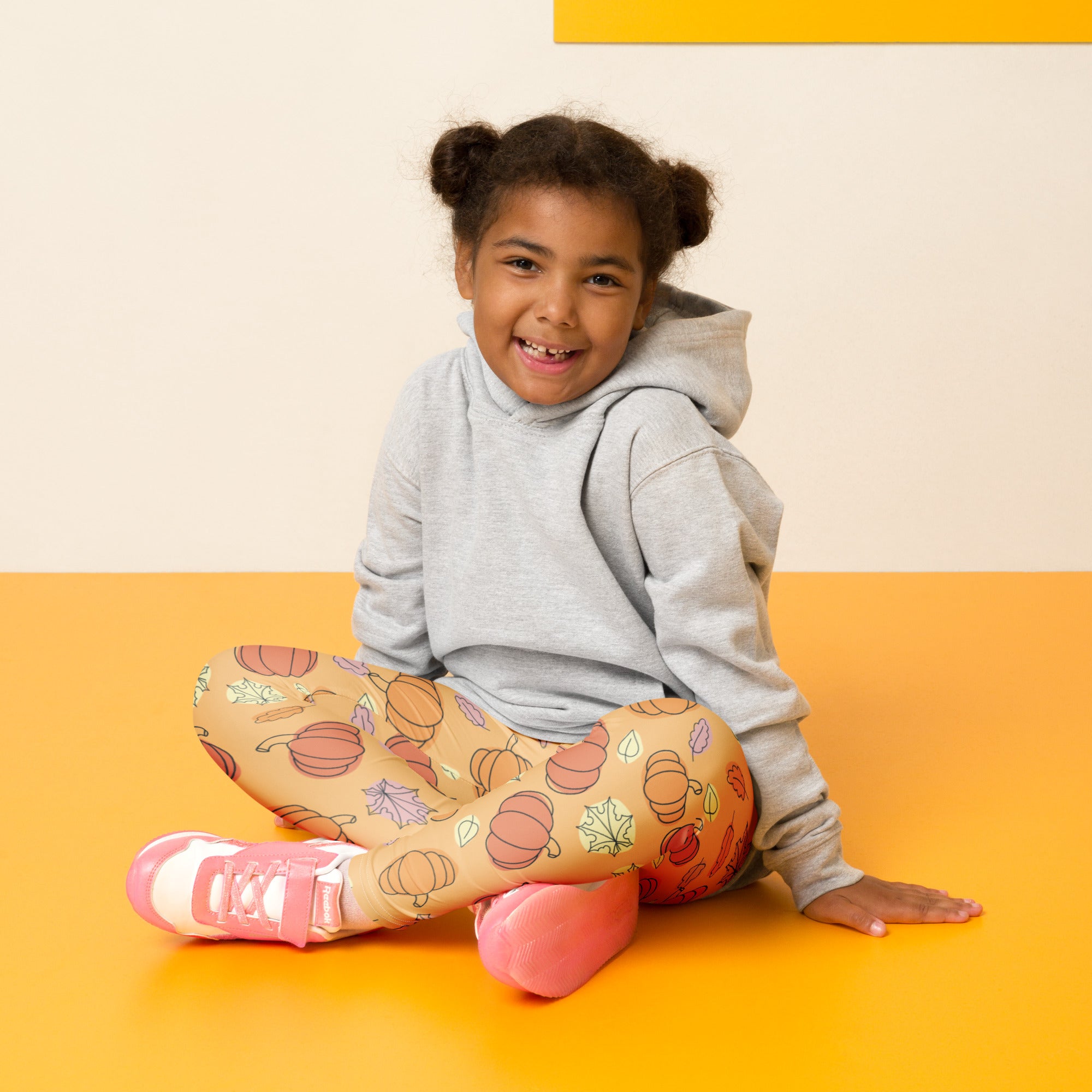 A child wearing buttery soft kids pumpkin patch leggings in beige with orange pumpkin and leaf patterns and pink sneakers