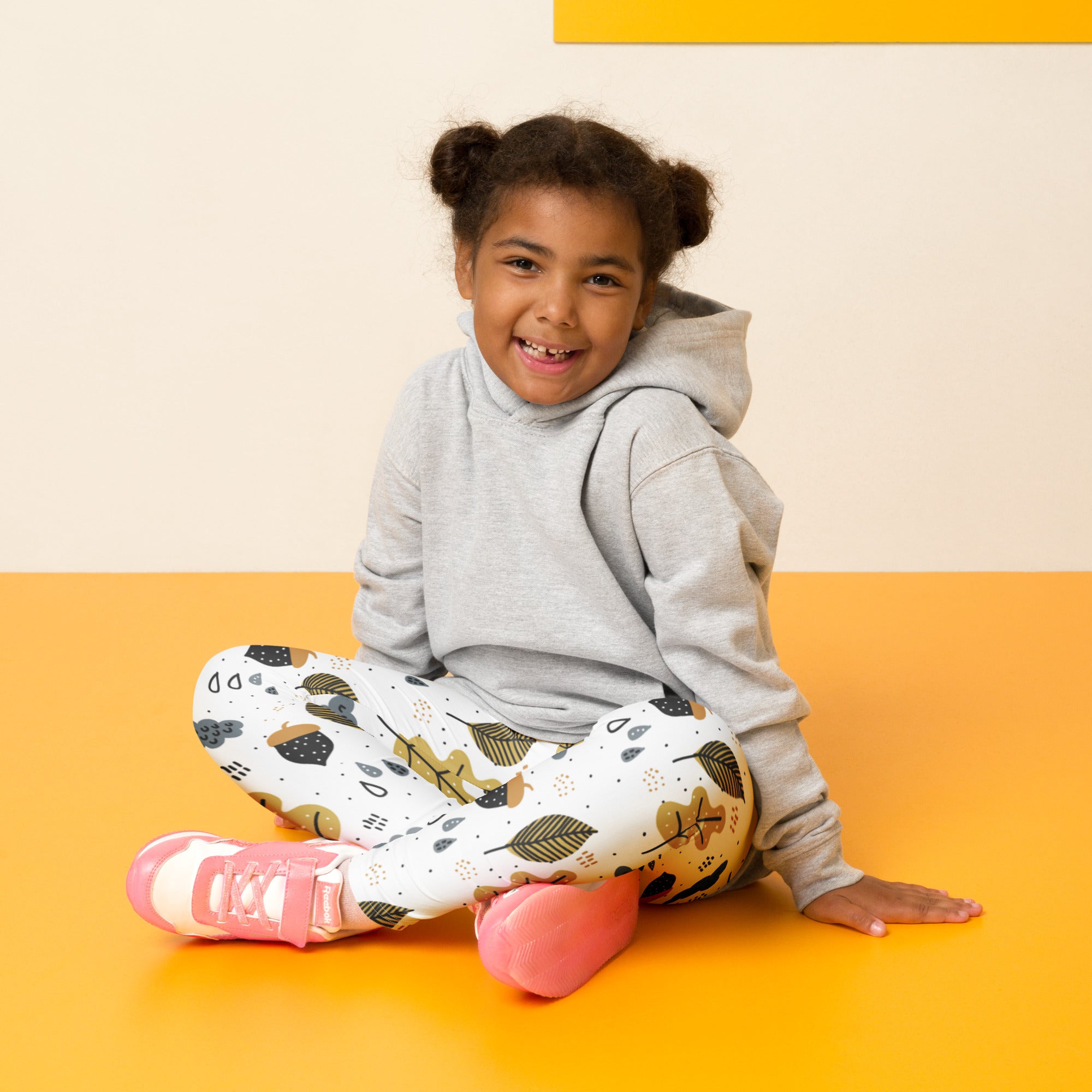 A child wearing buttery soft white leggings with autumn leaf patterns sits on an orange floor in fall season attire