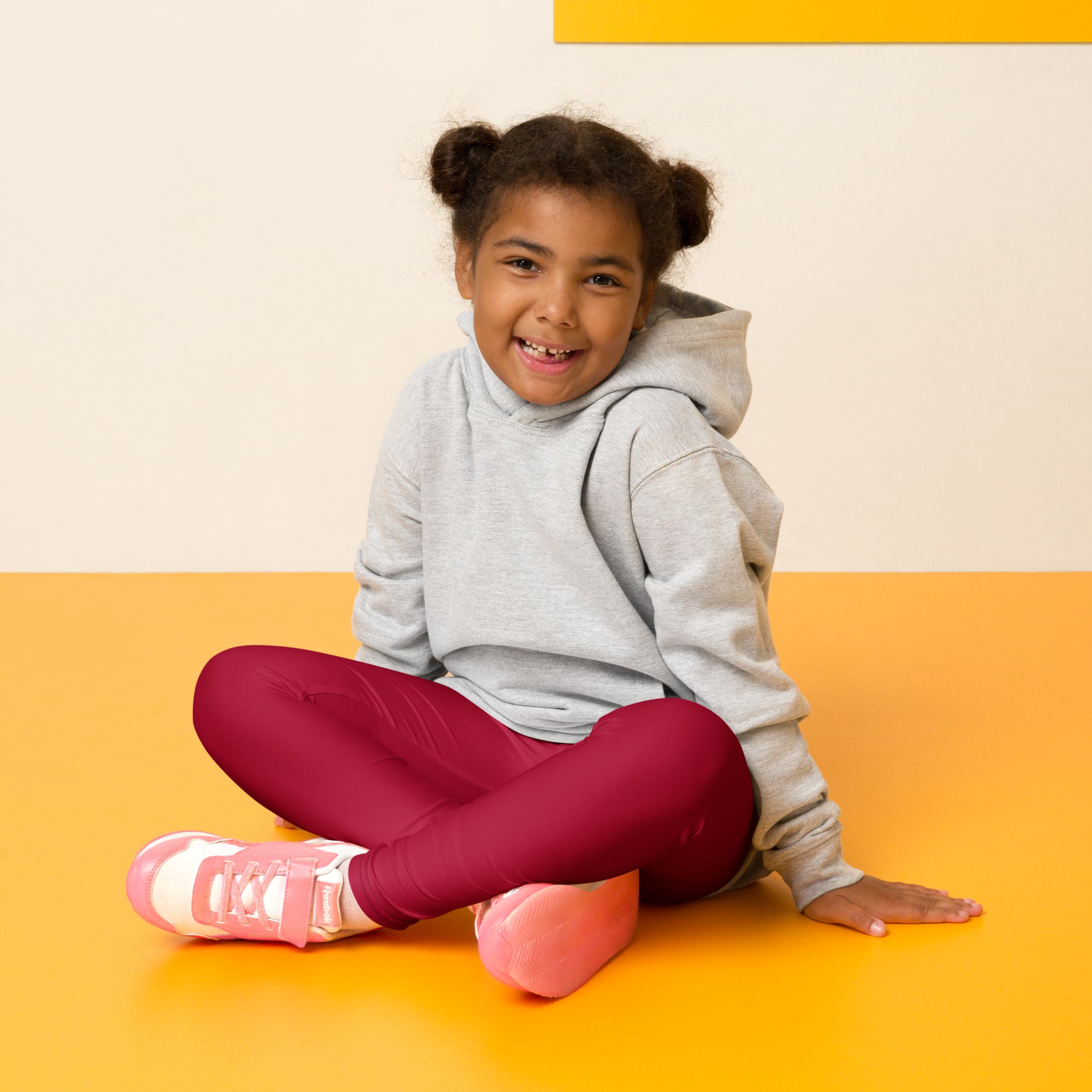 A child sits cross-legged on an orange floor wearing kids dark red buttery soft leggings and a gray hoodie
