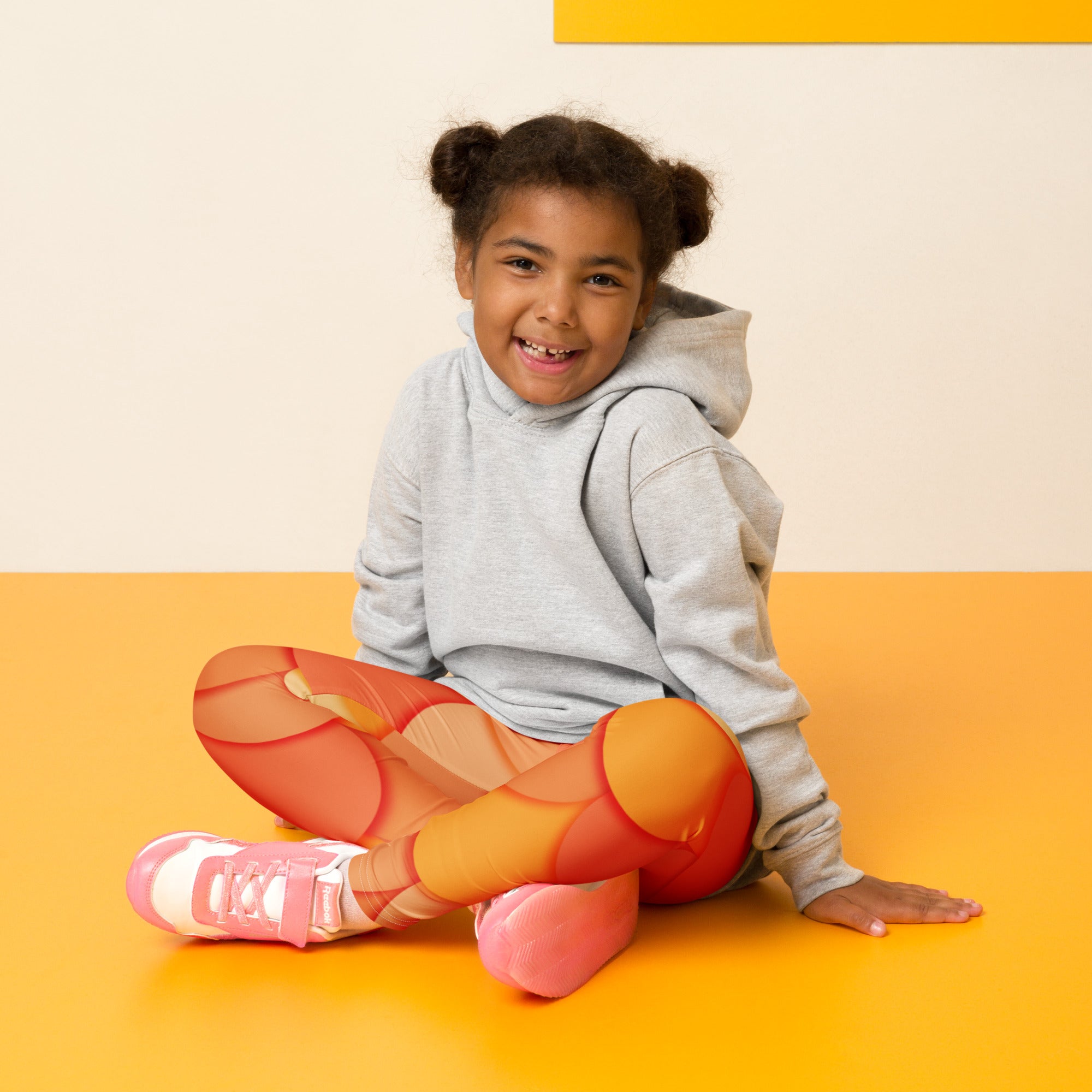A young girl wearing girls orange mermaid buttery soft leggings and a gray hoodie sitting cross-legged on an orange floor