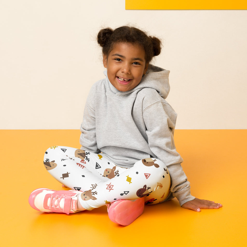 A child wearing gray hoodie sits on an orange floor wearing white buttery soft kids reindeer leggings with brown prints