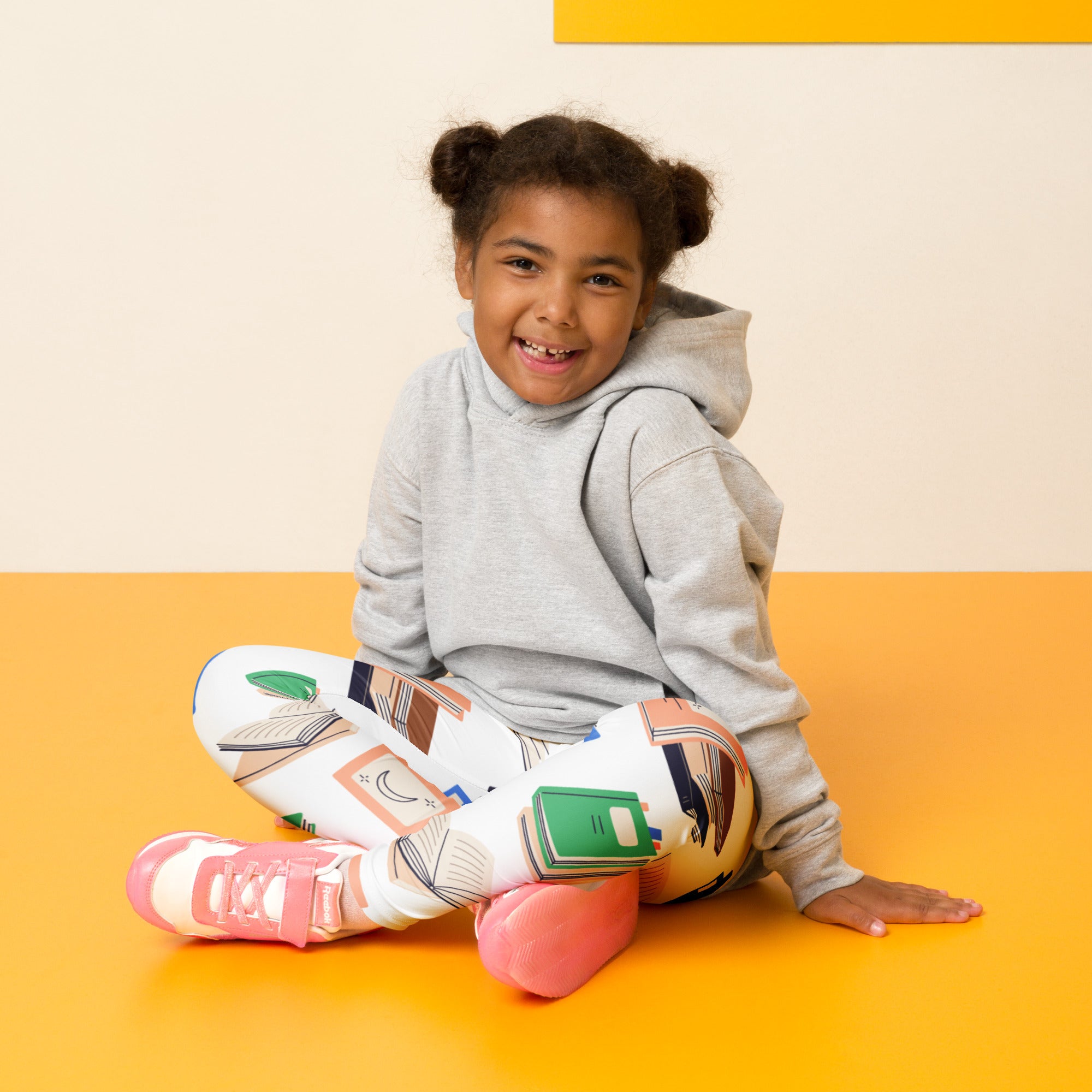 A child wearing kids book print buttery soft white leggings with colorful prints sitting on an orange floor