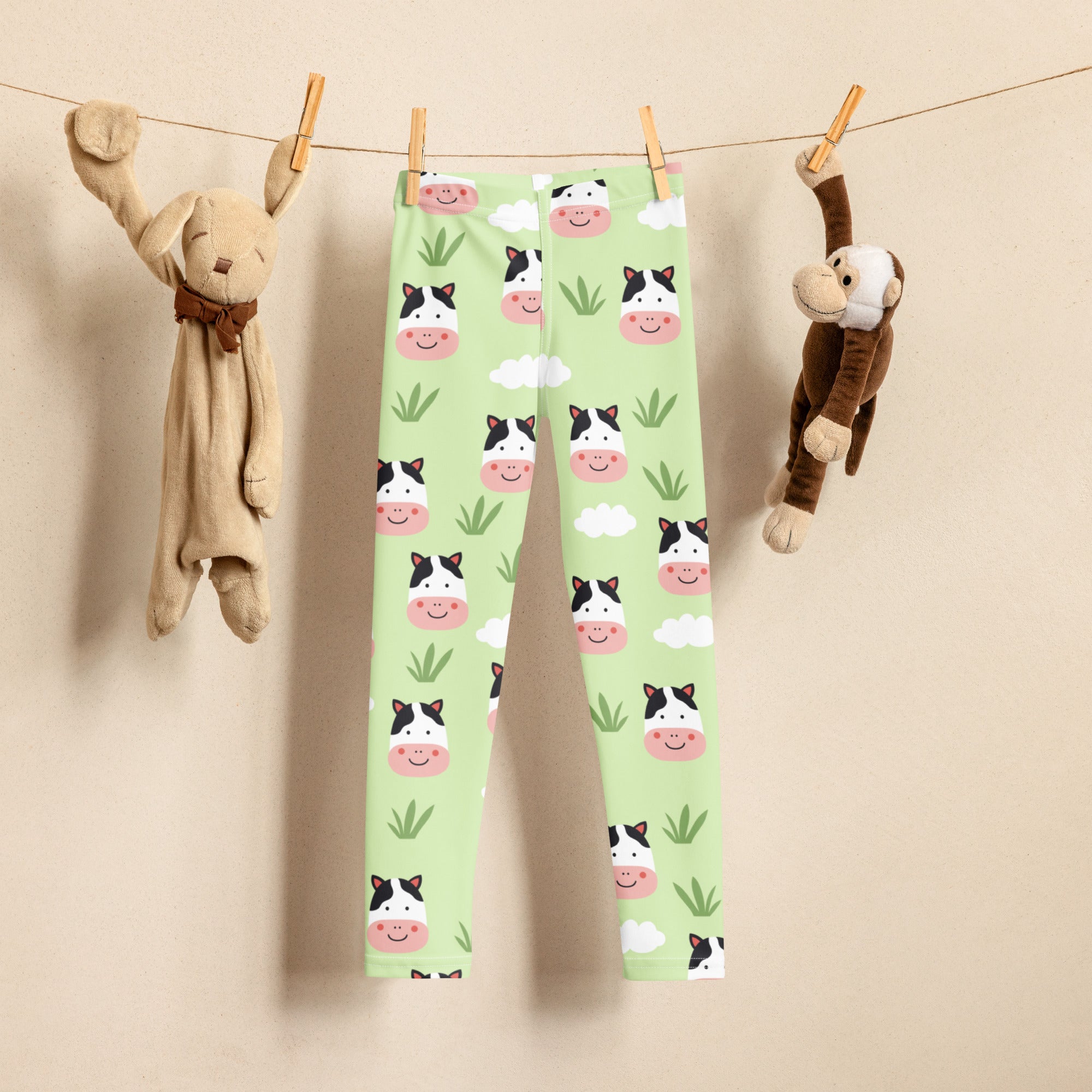 Light green kids buttery soft leggings with playful cartoon cow faces and grass pattern hanging on a clothesline