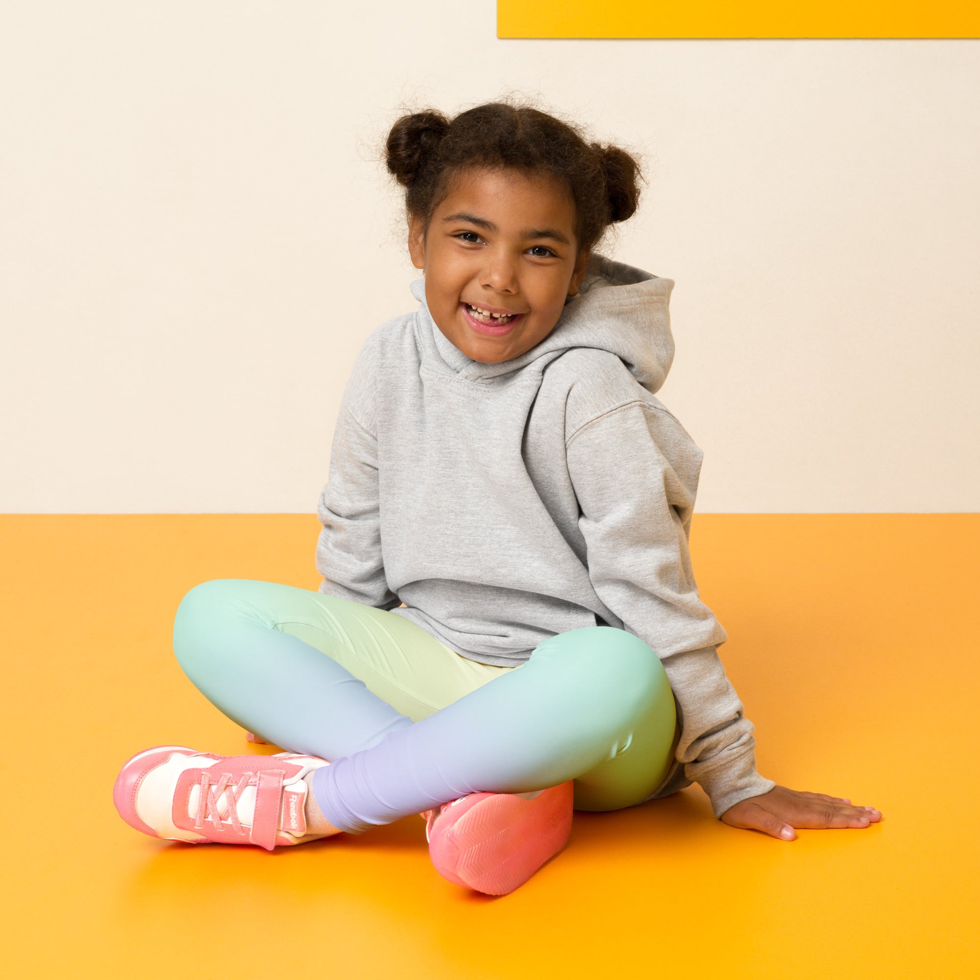 A child wearing girls pastel rainbow buttery soft leggings in a colorful gradient with pink sneakers and gray hoodie