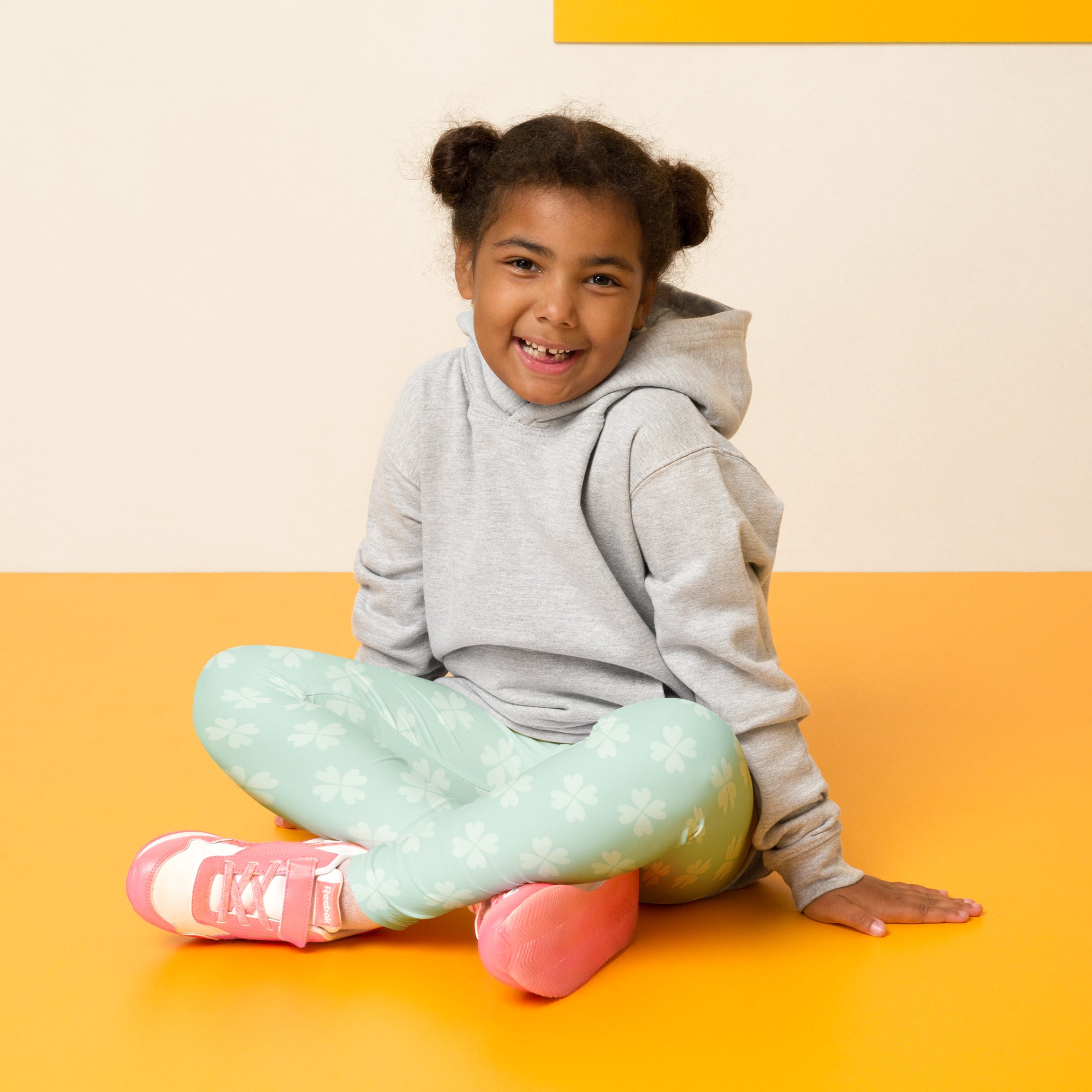 A young girl sits cross-legged wearing girls mint shamrock buttery soft leggings with white floral patterns on orange floor
