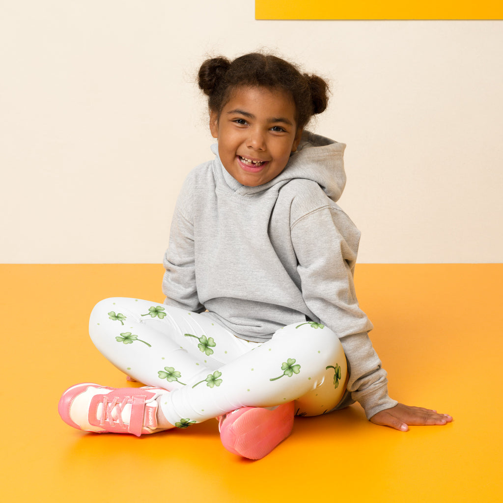 A young girl sits cross-legged wearing girls minimalist st patricks day clover buttery soft white leggings with green shamrocks