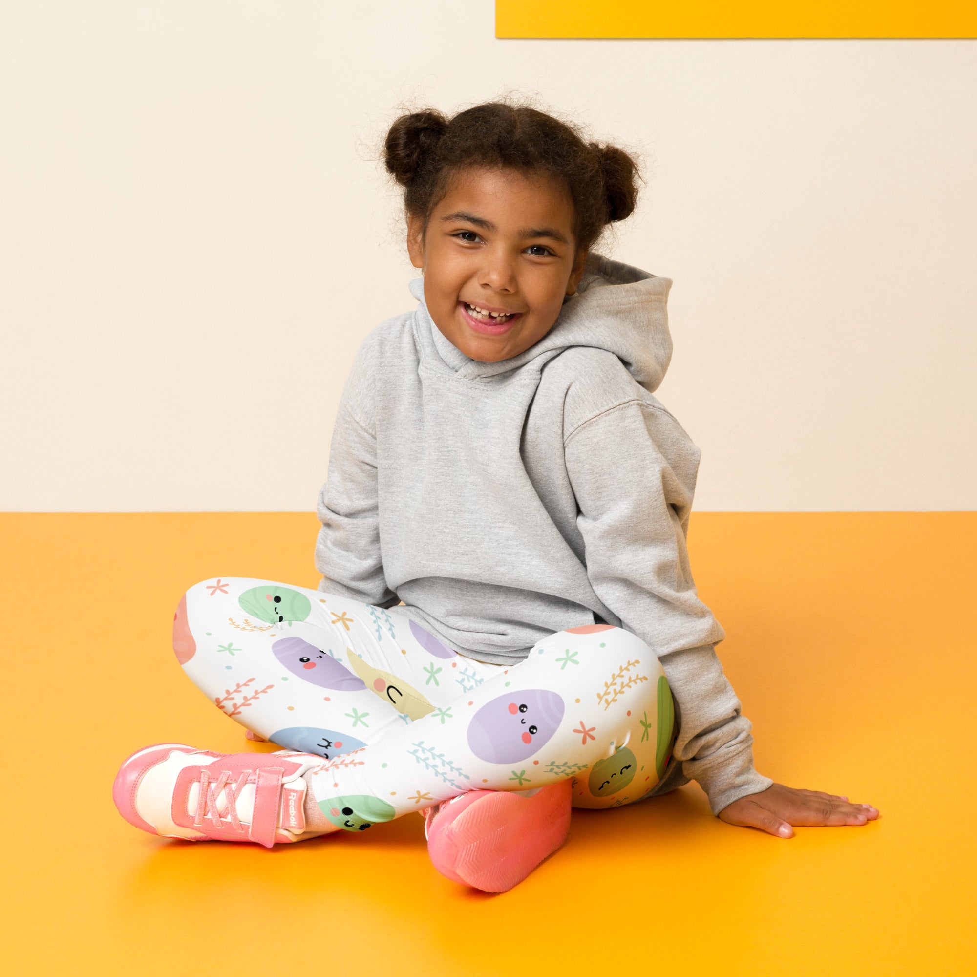 A child wearing gray hoodie and white buttery soft girls cute easter egg leggings with colorful prints and pink sneakers