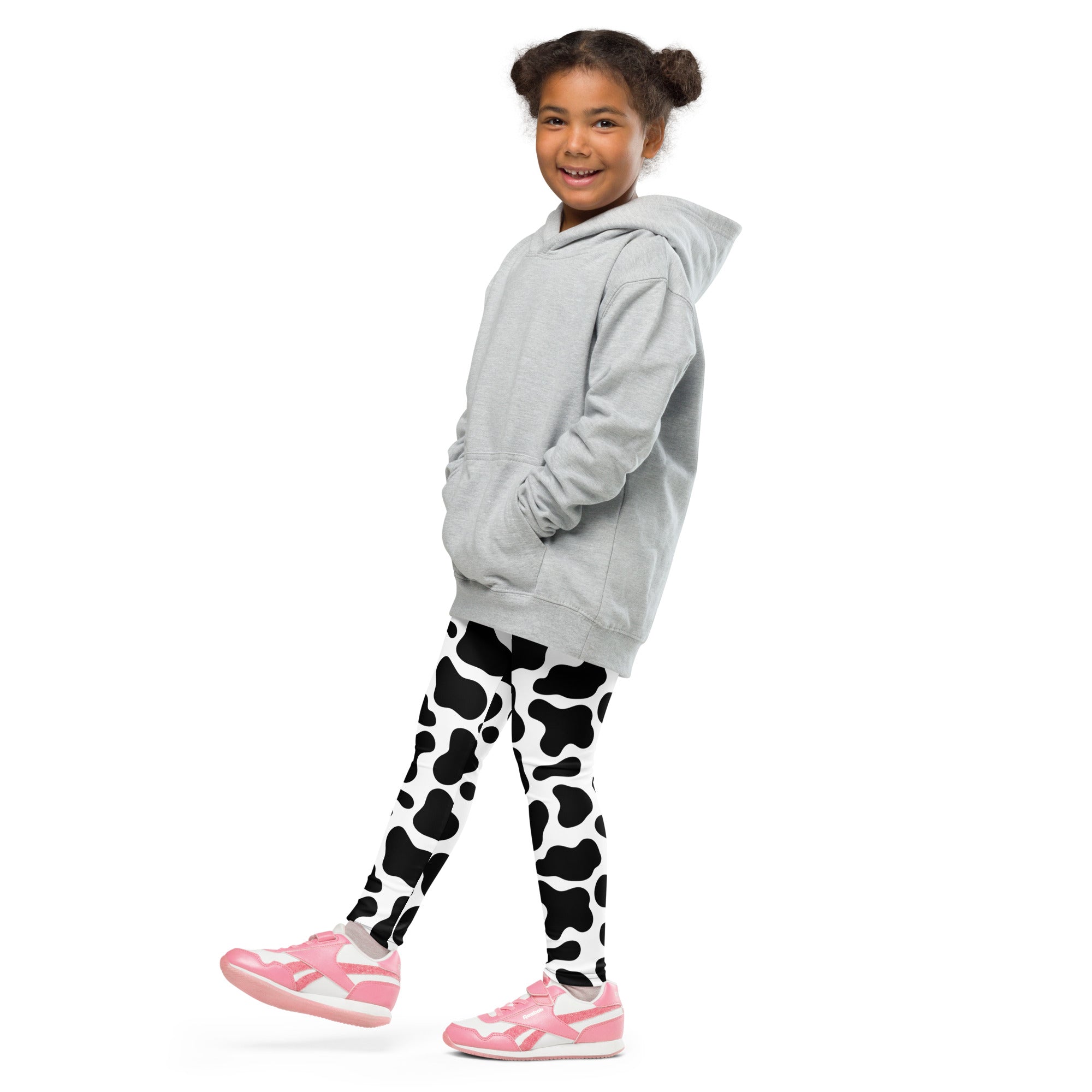 A young girl wearing kids cow print buttery soft black and white leggings with a light gray hooded sweatshirt and pink sneakers