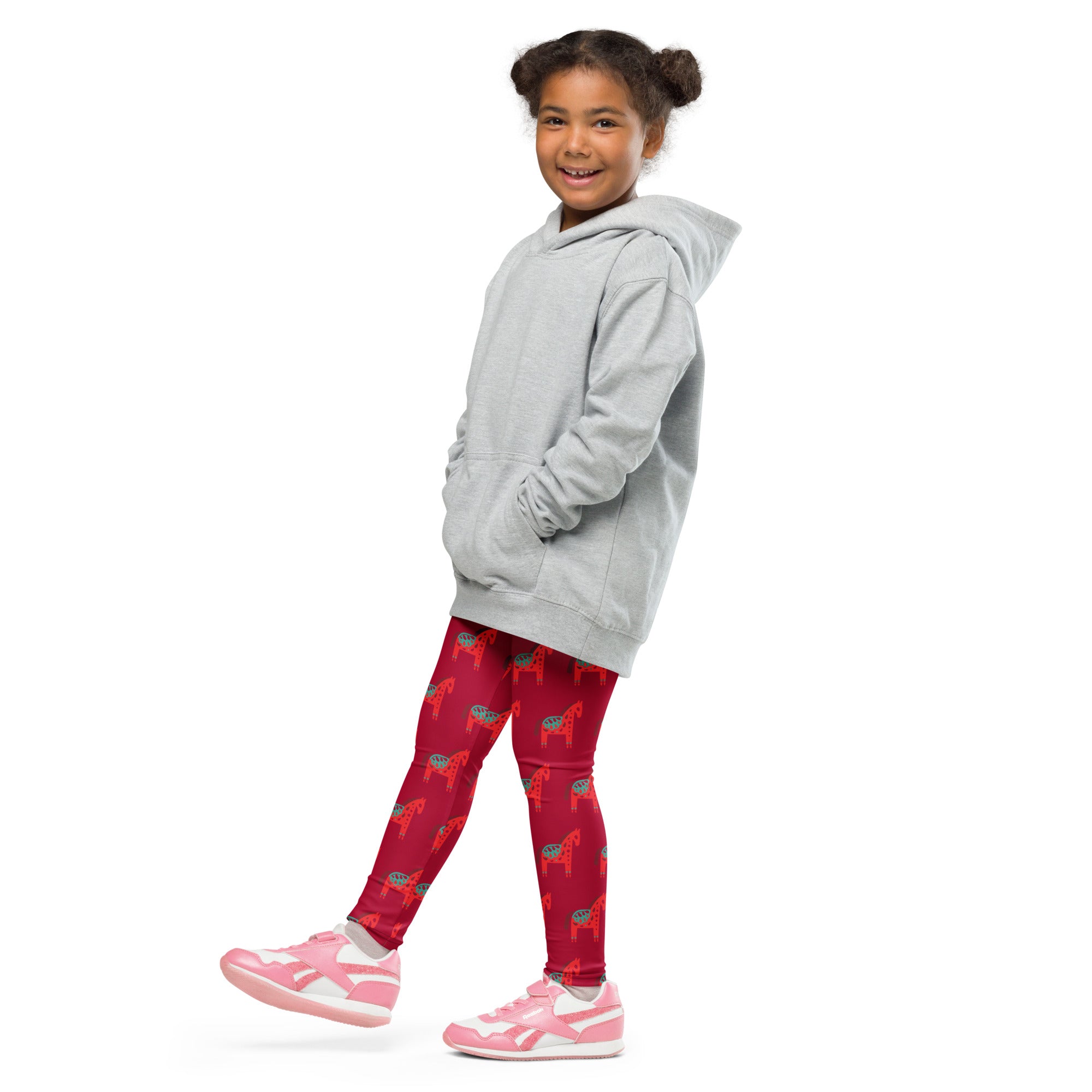 A young girl wearing buttery soft girls scandi horse Christmas leggings in red with horse patterns and a gray hoodie