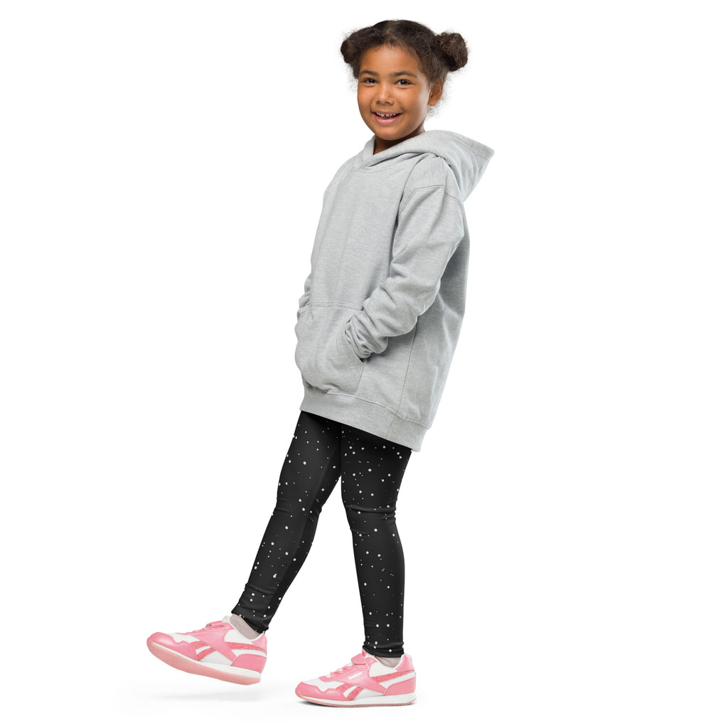 A young girl wearing kids black starry night buttery soft leggings with white stars, paired with a gray hoodie and pink sneakers