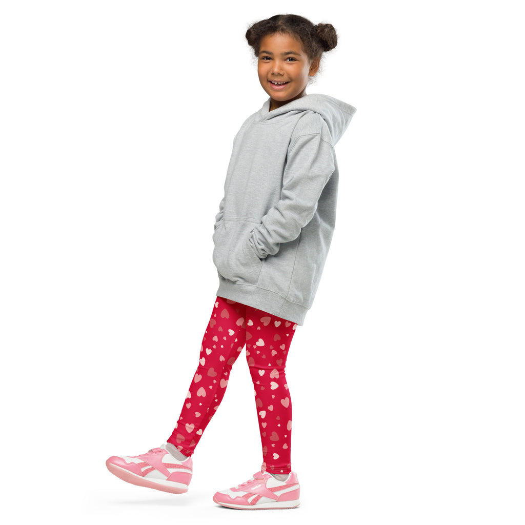 A young girl wearing girls red heart print buttery soft leggings in bright red with scattered pink hearts and pink sneakers