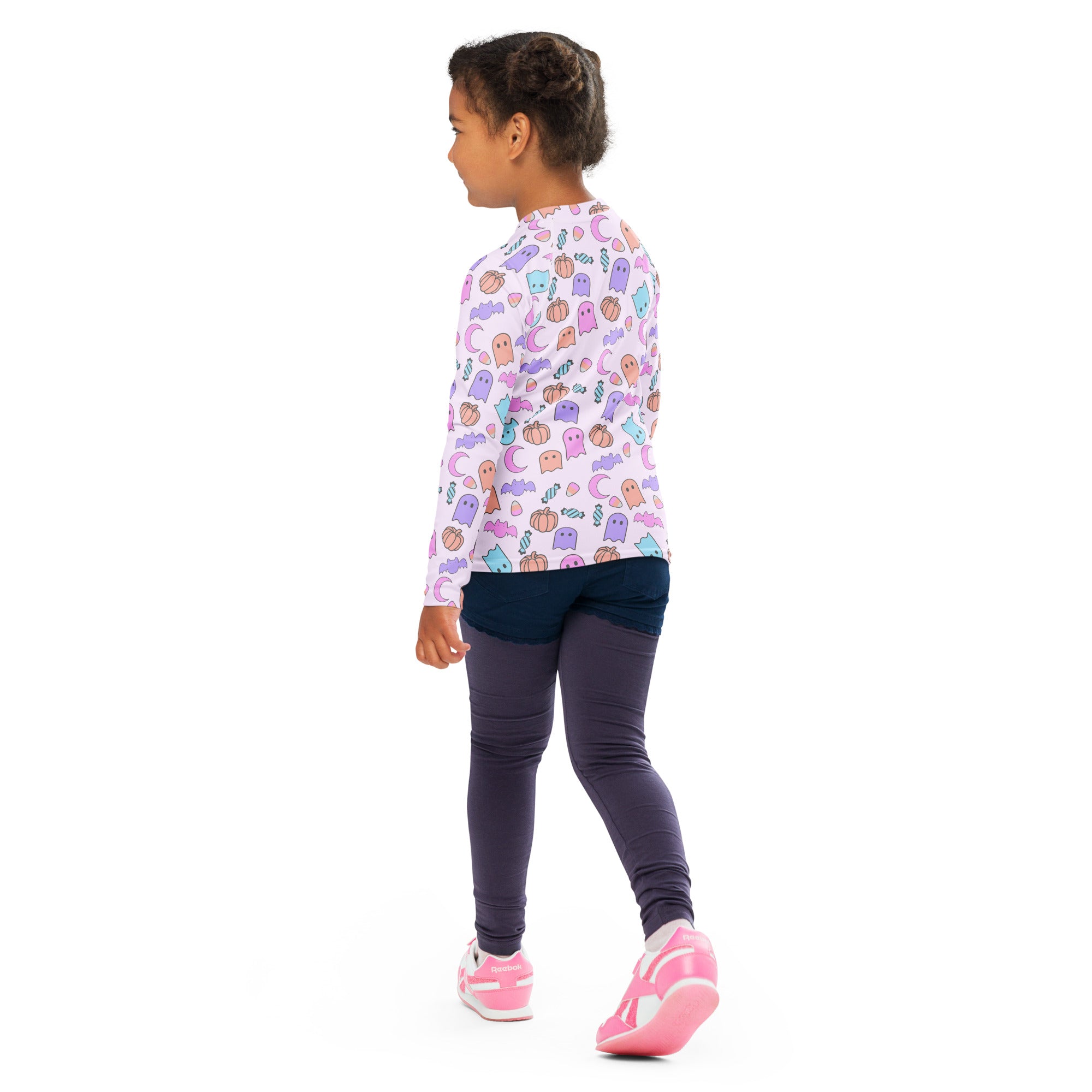 A girl wearing a white long sleeve halloween rash guard swim shirt with pastel ghost pumpkin and moon pattern upf 50+