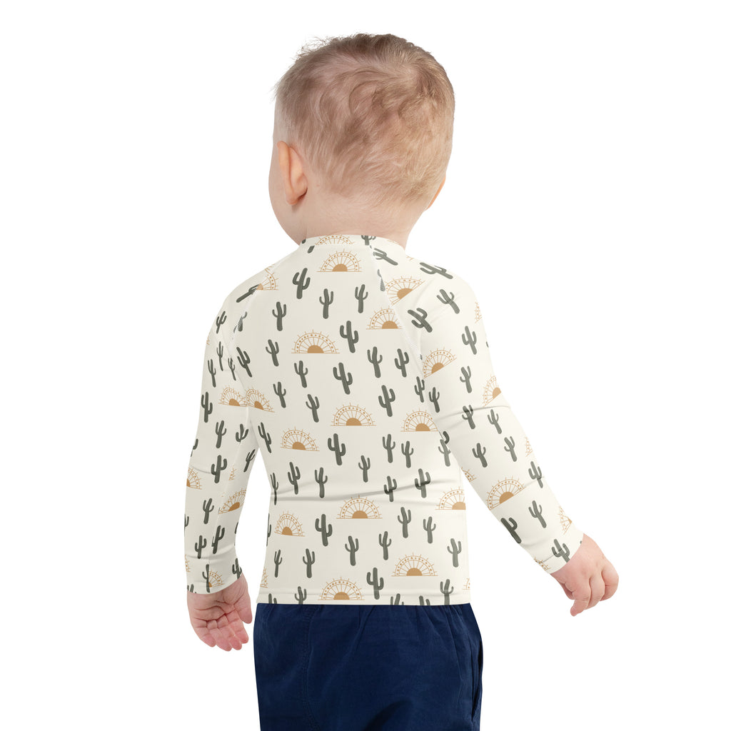 A child wearing a cream kids cactus desert sunrise long sleeve rash guard swim shirt with UPF 50 and black cactus gold sun pattern