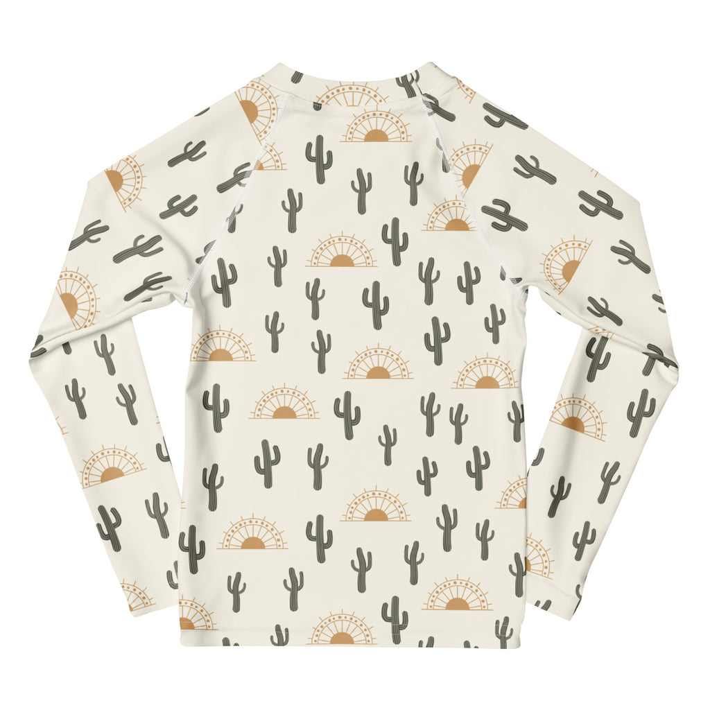 Cream-colored kids long sleeve rash guard with repeating black cacti and tan sunburst pattern upf 50+ swim shirt