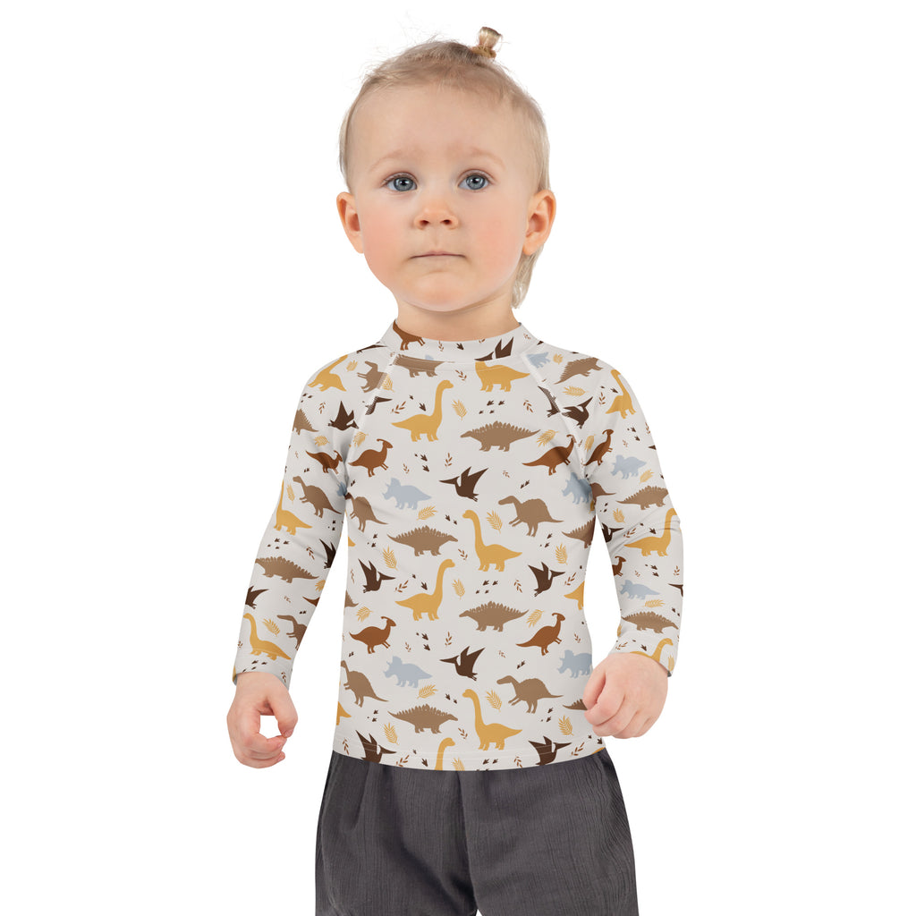 A child wearing a cream longsleeve kids dinosaur rash guard swim shirt with brown gold dinosaur print and UPF 50 plus protection