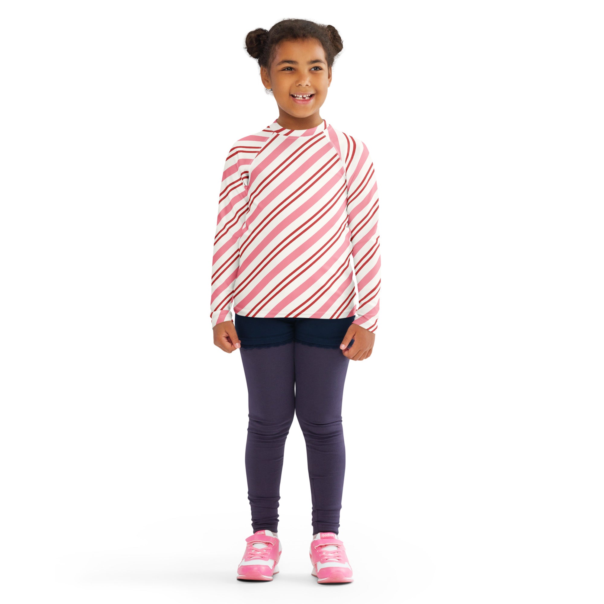 A girl wearing a kids pink peppermint stripe Christmas long sleeve rash guard swim shirt with UPF 50+ and purple leggings