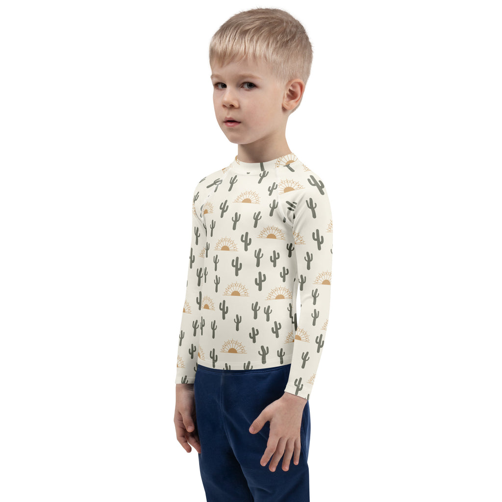 A child wearing a cream kids cactus desert sunrise long sleeve rash guard swim shirt with UPF 50 and black cacti pattern