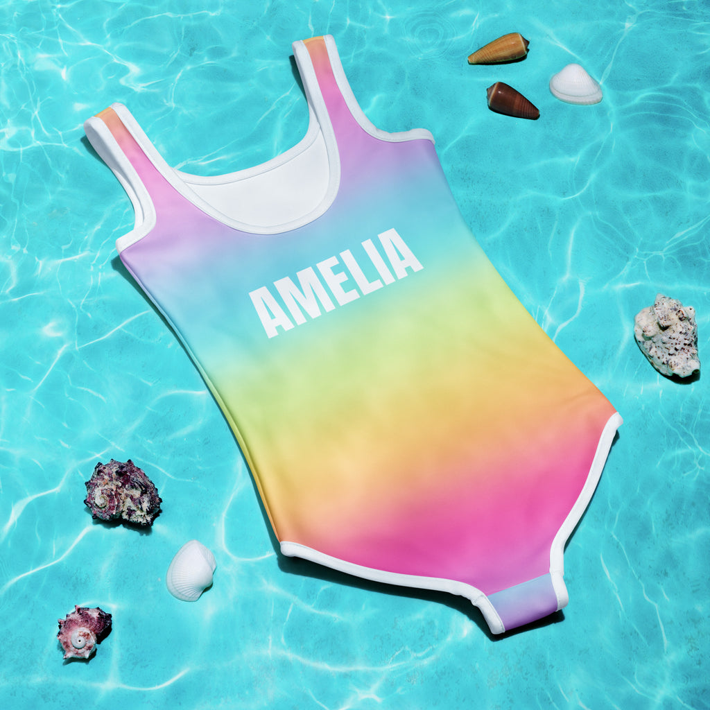 A girls bright rainbow gradient one-piece swimsuit with white trim and name amelia printed on chest in clear blue water