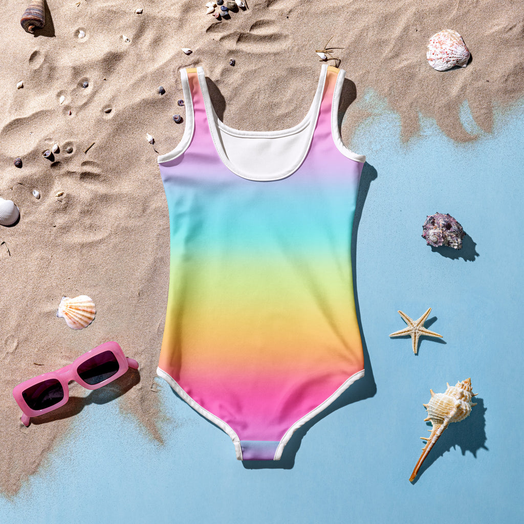Girls bright rainbow buttery soft one-piece swimsuit with white trim scoop neckline laid on sandy beach with upf 50+