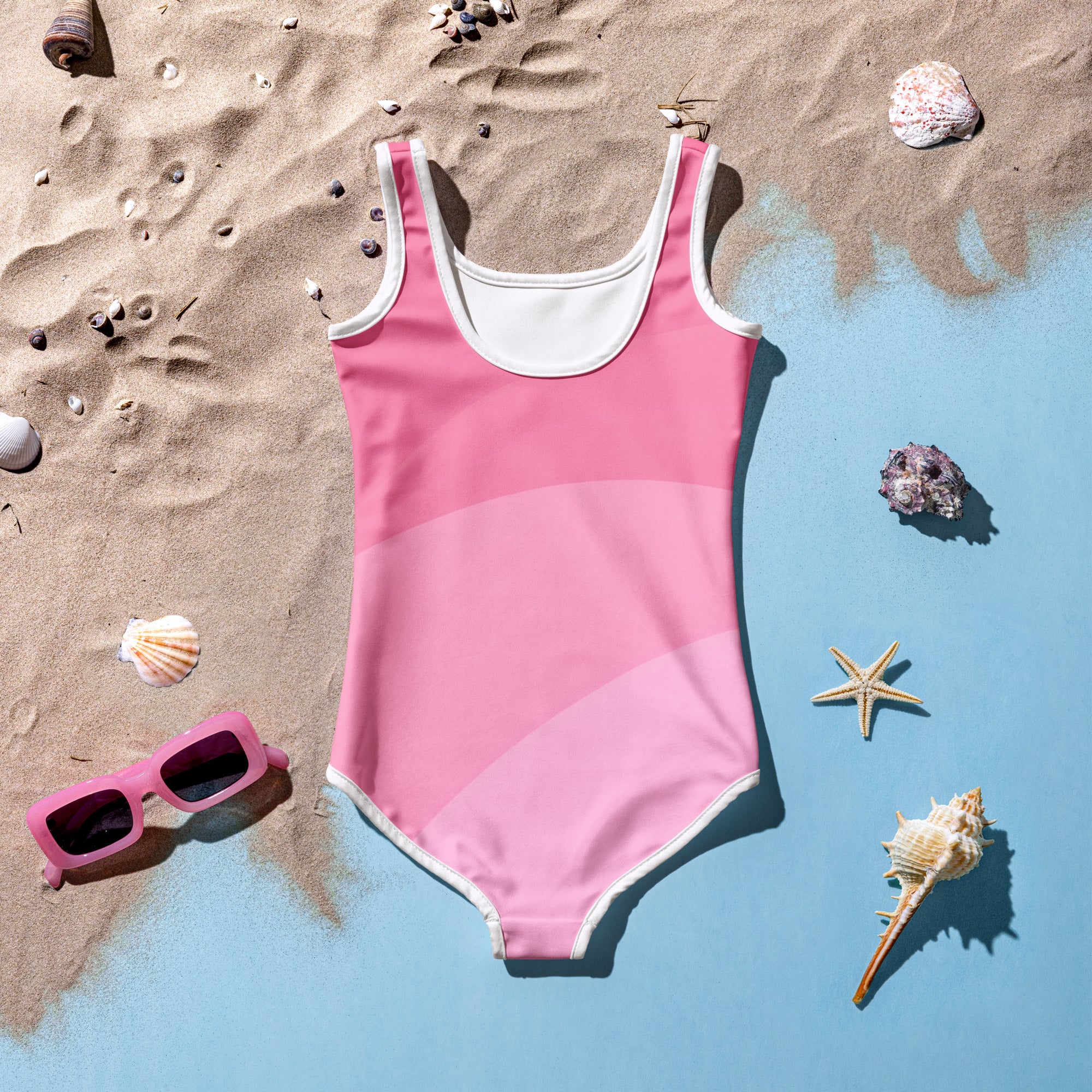 Girls wavy pink swimsuit one-piece with white trim and subtle gradient laid on sand beside seashells