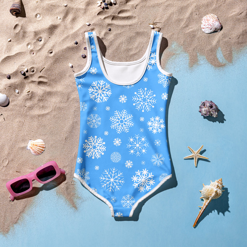 A vibrant blue girls snowflake Christmas one-piece swimsuit with white trim laid on sandy beach near water edge