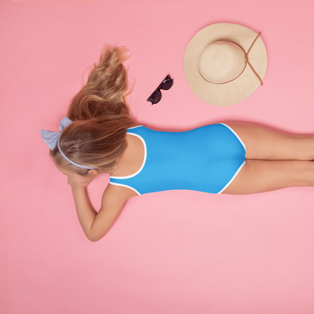 A woman lies on her stomach wearing a girls sky blue buttery soft one-piece swimsuit with white trim and UPF 50+ protection