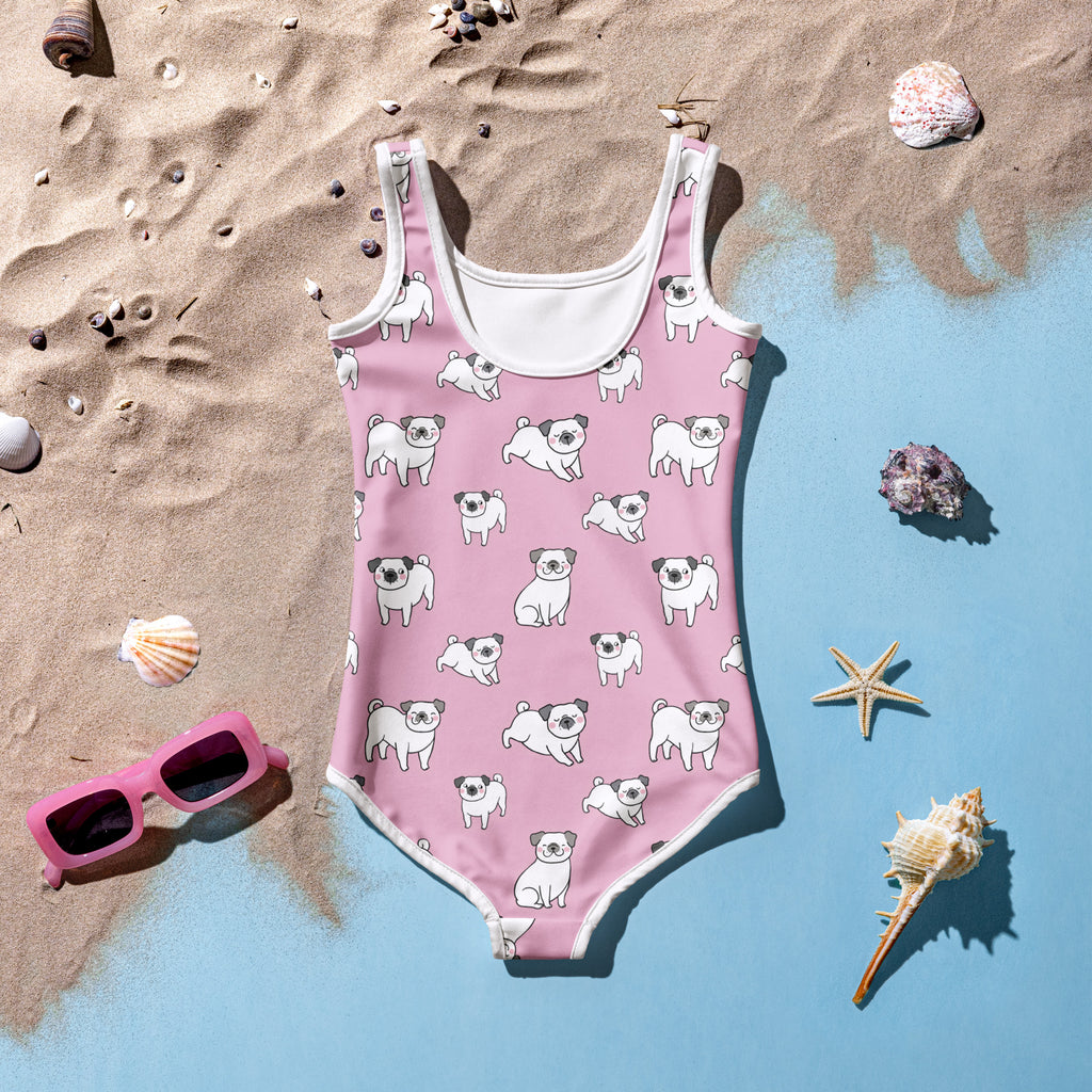 Girls pink swimsuit with dog pattern on a sandy and blue gradient background with sunglasses and shells.
