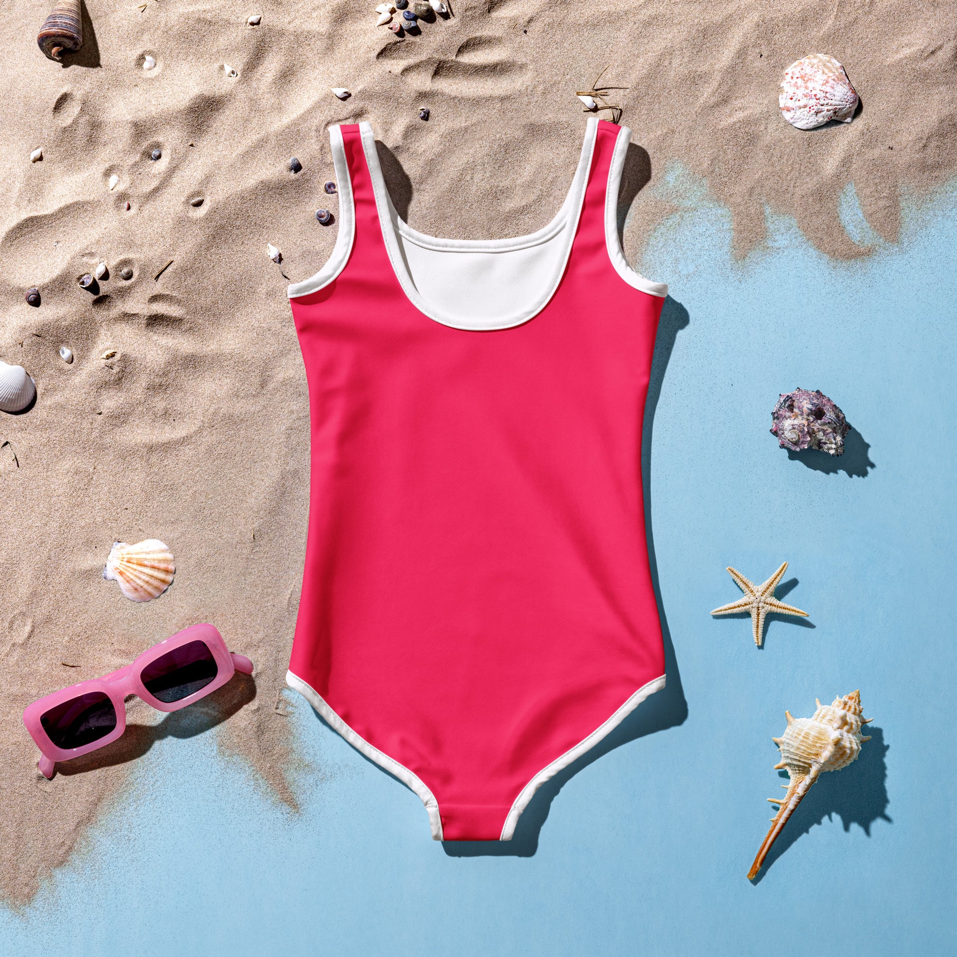 Crimsom pink swimsuit with white trim on a sandy surface with shells and sunglasses.