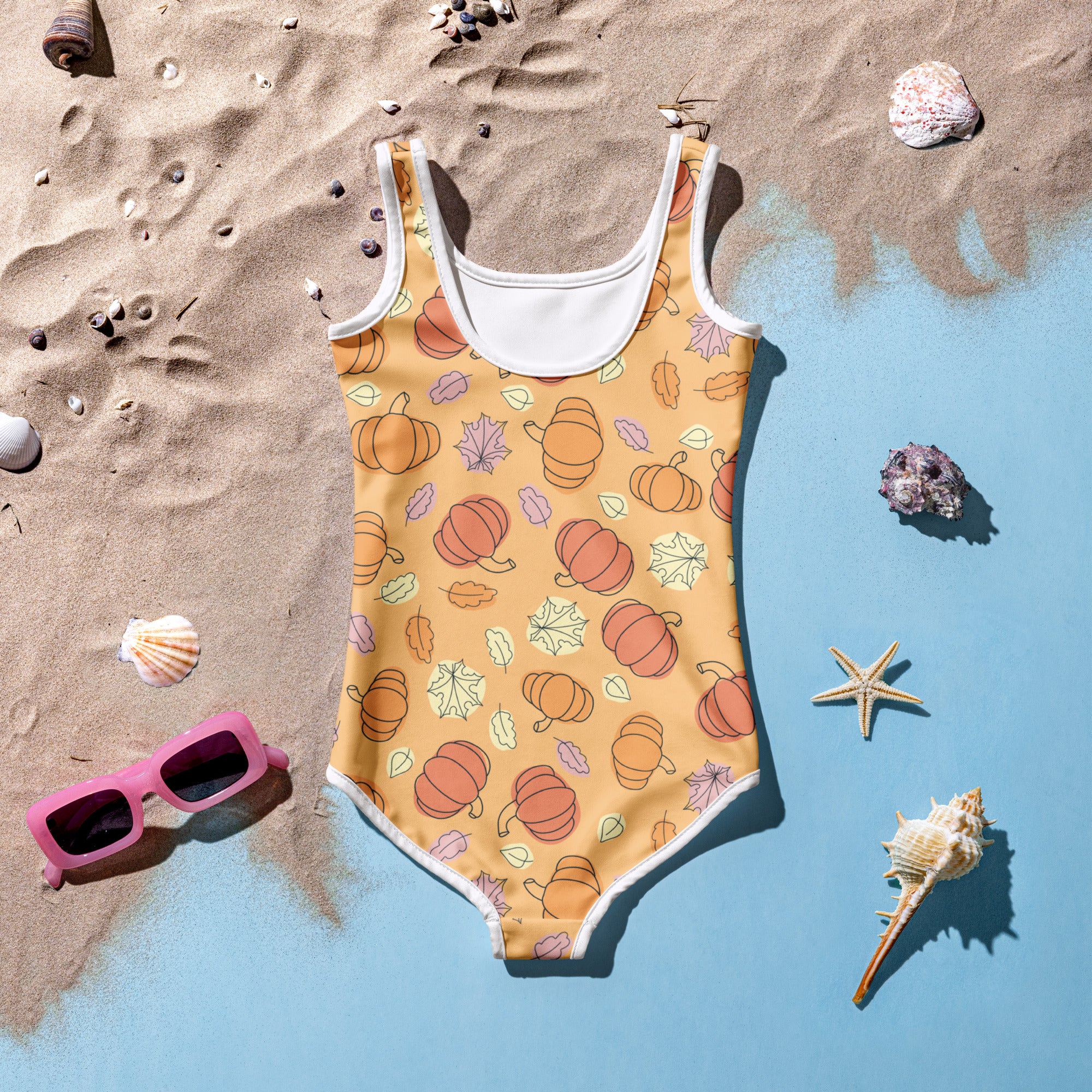 One-piece swimsuit with pumpkin pattern on a sandy surface with shells and sunglasses.