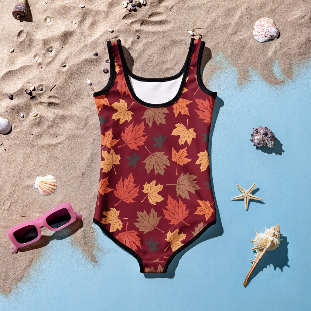 One-piece swimsuit with autumn leaf pattern on a sandy and blue gradient background with shells and sunglasses.