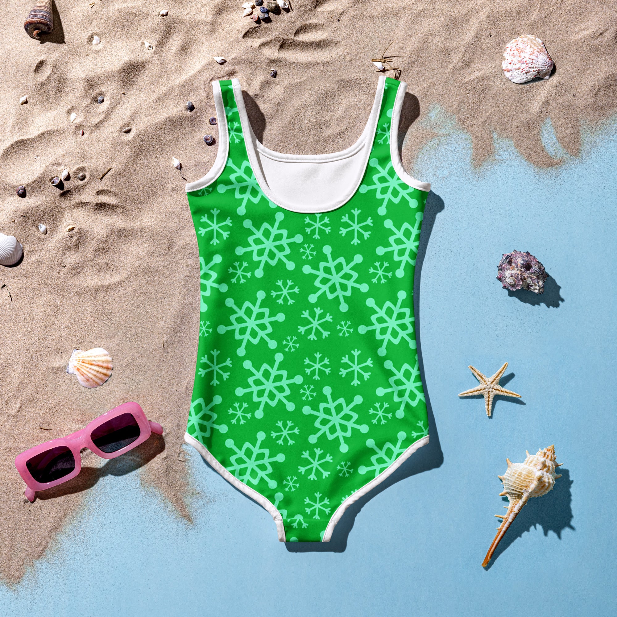 Girls green one-piece buttery soft swimsuit with white trim and mint snowflake pattern laid on sandy beach near water