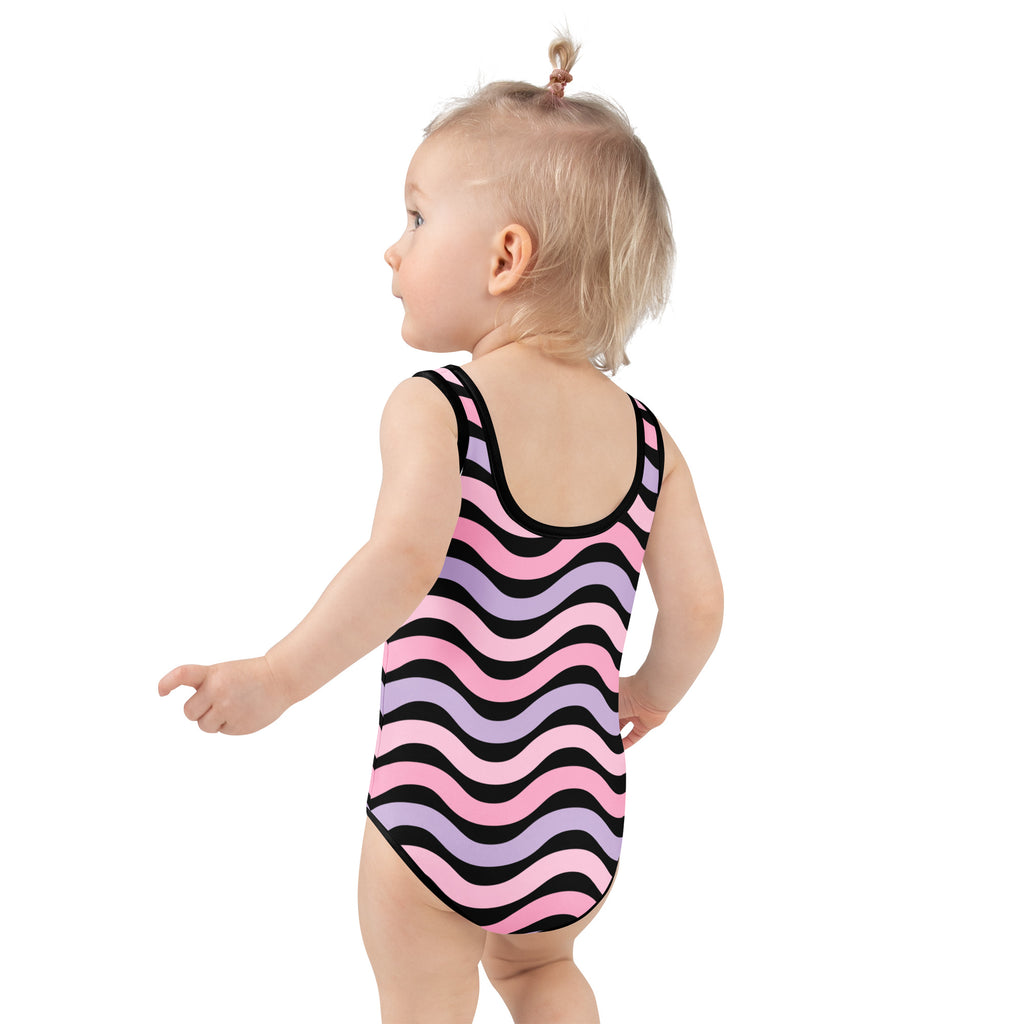 A toddler wearing a girls pink and lavender wavy stripe buttery soft swimsuit with UPF 50 plus and double layered front