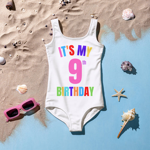 A vibrant white swimsuit is displayed on a sandy beach, featuring colorful text that reads ITS MY 9th BIRTHDAY. Surrounding the swimsuit are seashells and a starfish, along with a pair of pink sunglasses, creating a festive beach scene.