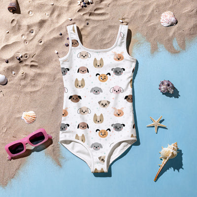 White kids swimsuit with cute animal faces print, displayed on sand with seashells and pink sunglasses